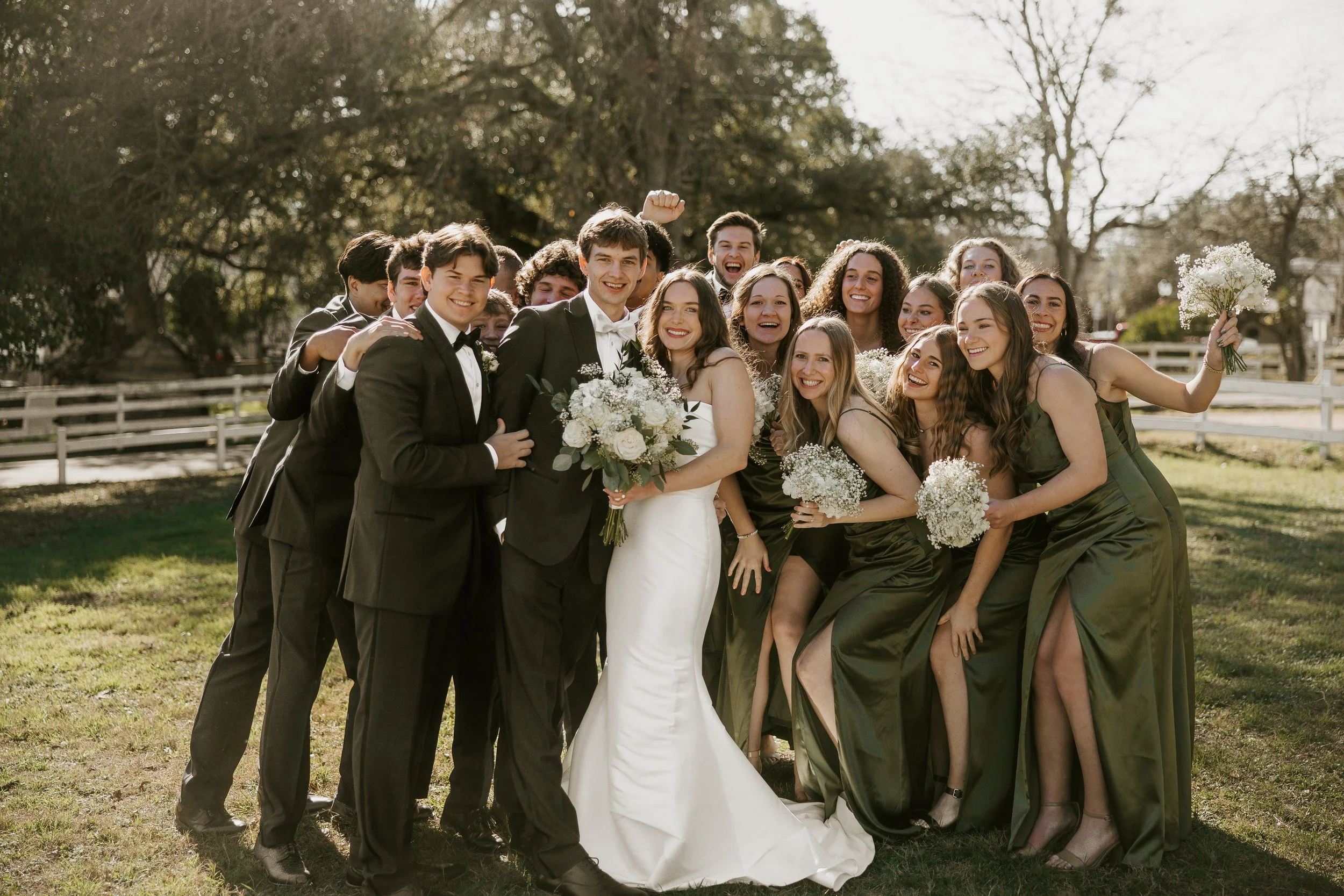 A group of people in formal attire, including a bride in a white wedding dress and groom in a black tuxedo, posing together outdoors on a grassy area with trees and a white fence in the background.
