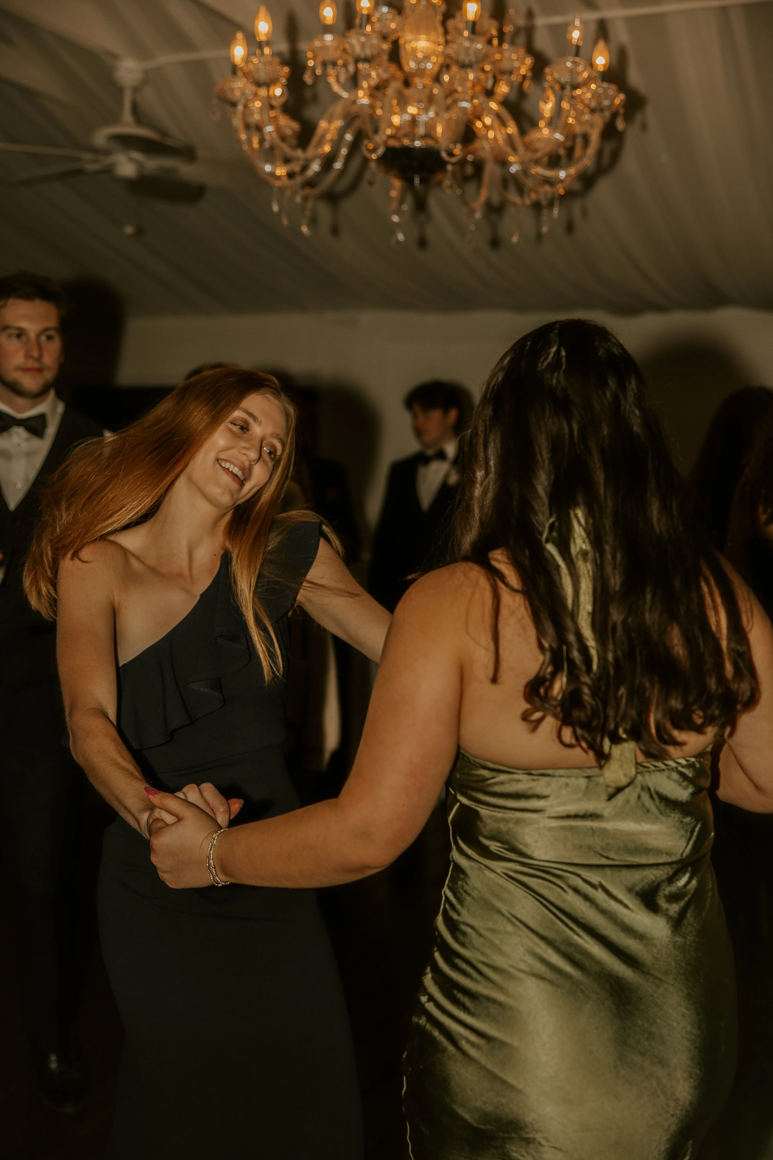 Two women dancing and holding hands at a formal event with a chandelier overhead, while men in tuxedos observe in the background.