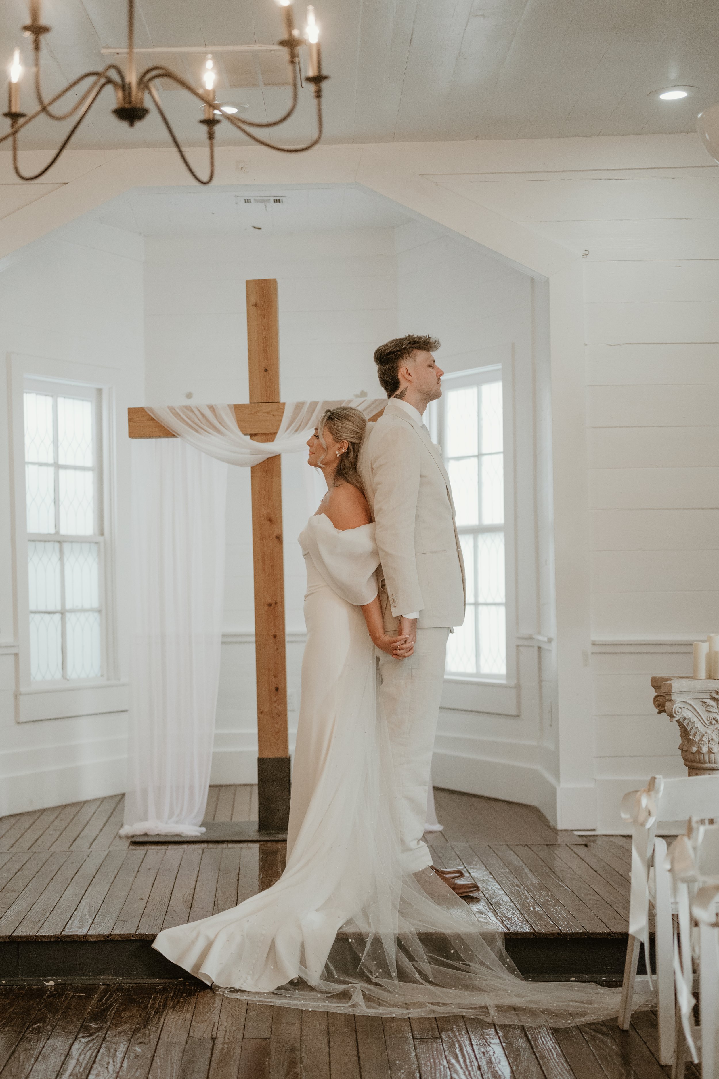 A bride and groom standing back-to-back holding hands in front of a wooden cross with white drapery in a bright, white room with windows.