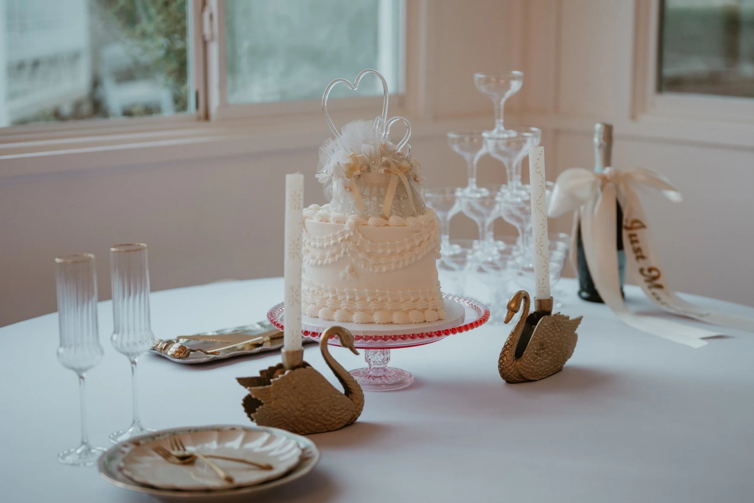 Elegant white wedding cake with heart decoration, surrounding by champagne glasses, candles, and gold swan decorations on a round table in front of a window.
