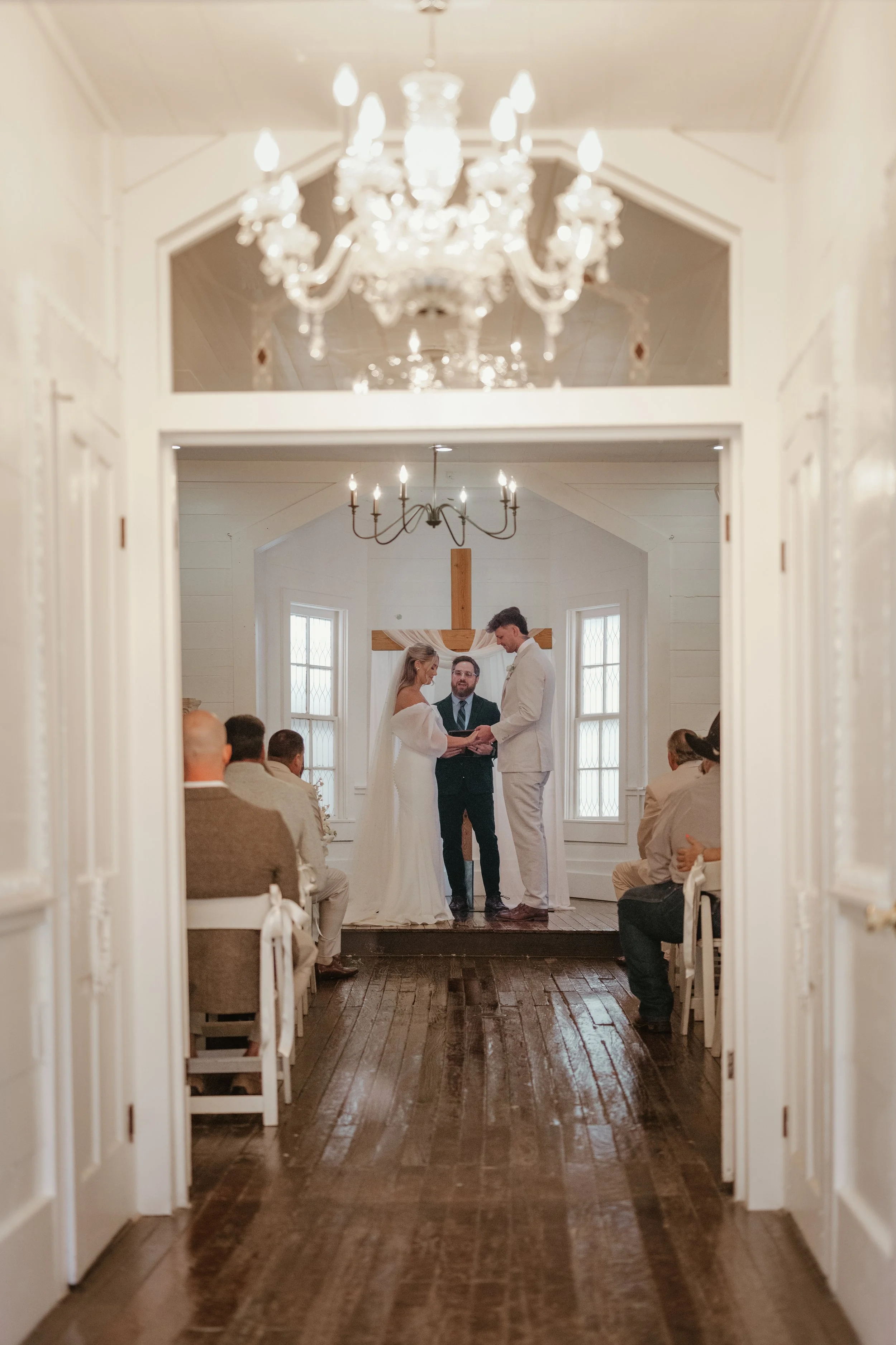 A wedding ceremony taking place in a small chapel, with the bride and groom exchanging vows in front of an officiant, all seen through a doorway. The chapel has white walls, tall windows, wooden floor, and chandeliers overhead.