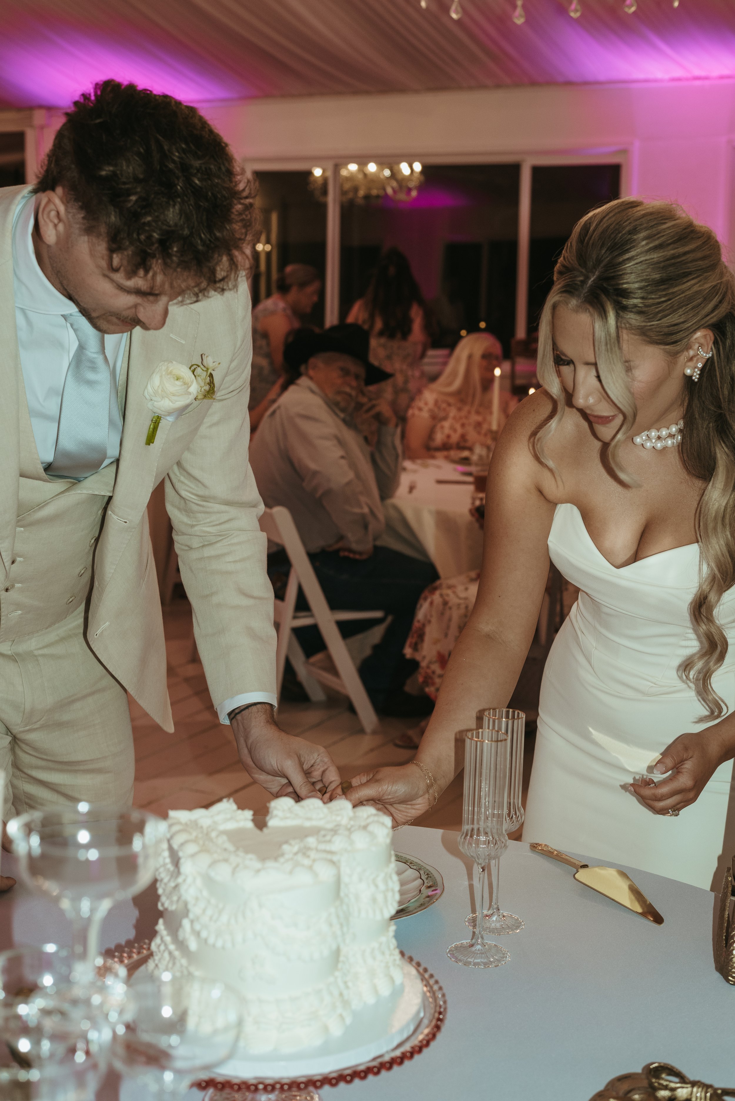 A bride and groom cutting a wedding cake at their reception, with guests seated at tables in the background.