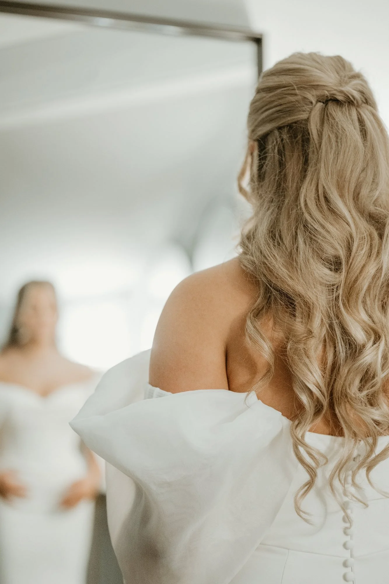 A bride with long, curly blonde hair looking at her reflection in a mirror, dressed in a white wedding gown.