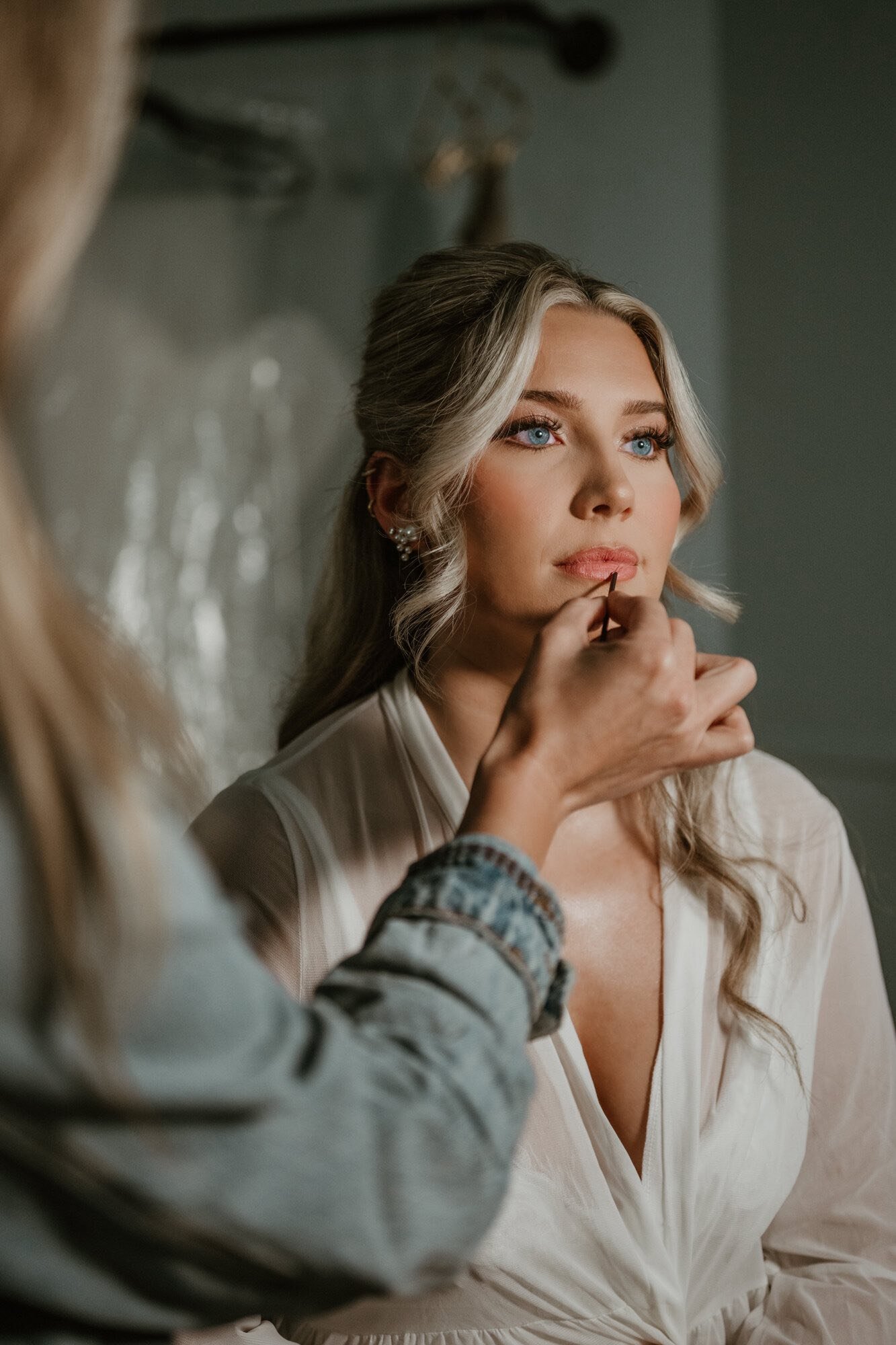 A woman with blue eyes and blonde hair is getting her makeup done by a makeup artist, who is applying lipstick to her lips.