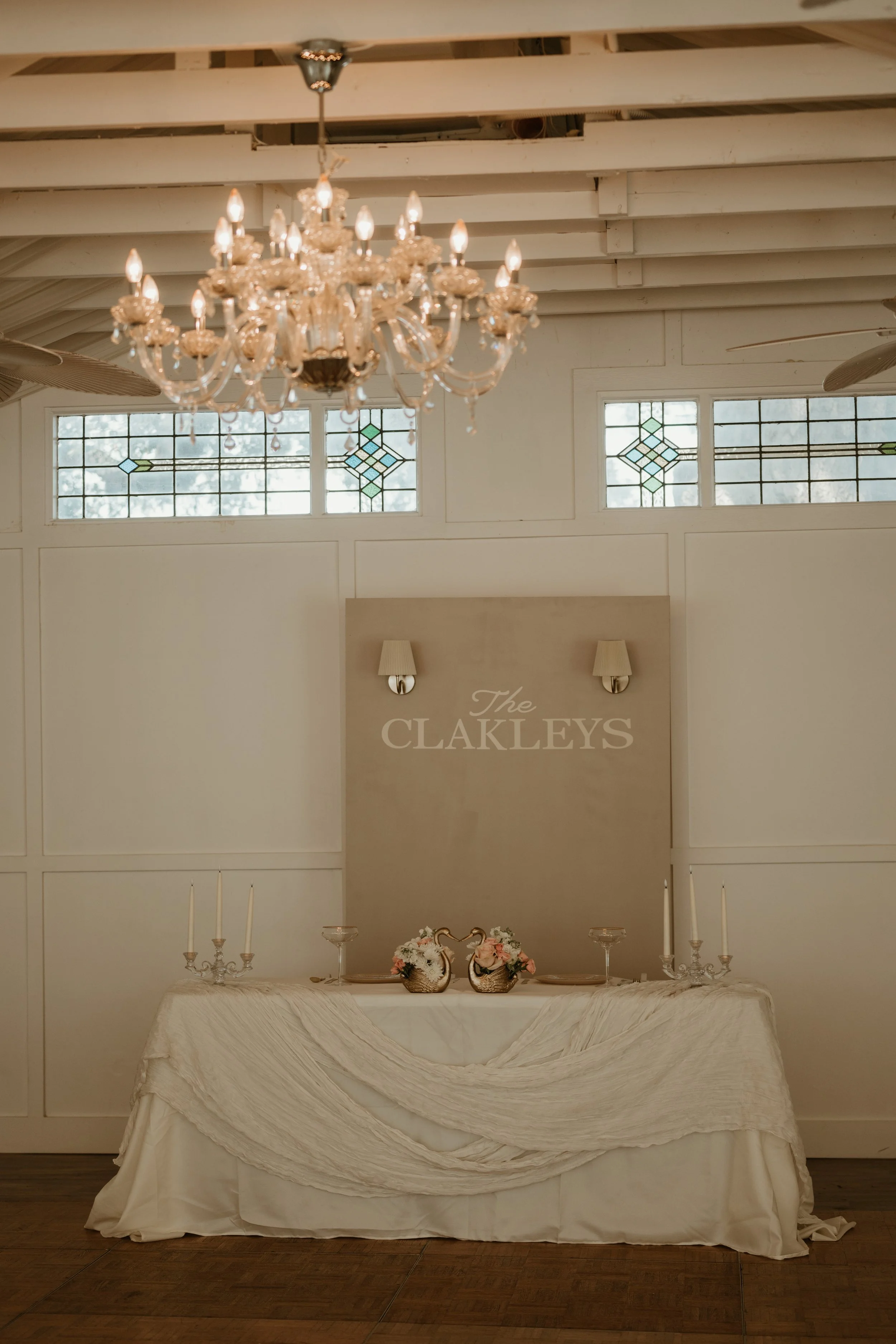 Elegant wedding reception table with floral arrangements, candelabras, and a backdrop with the text 'The Clakeys' in a sophisticated venue with stained glass windows and a chandelier.