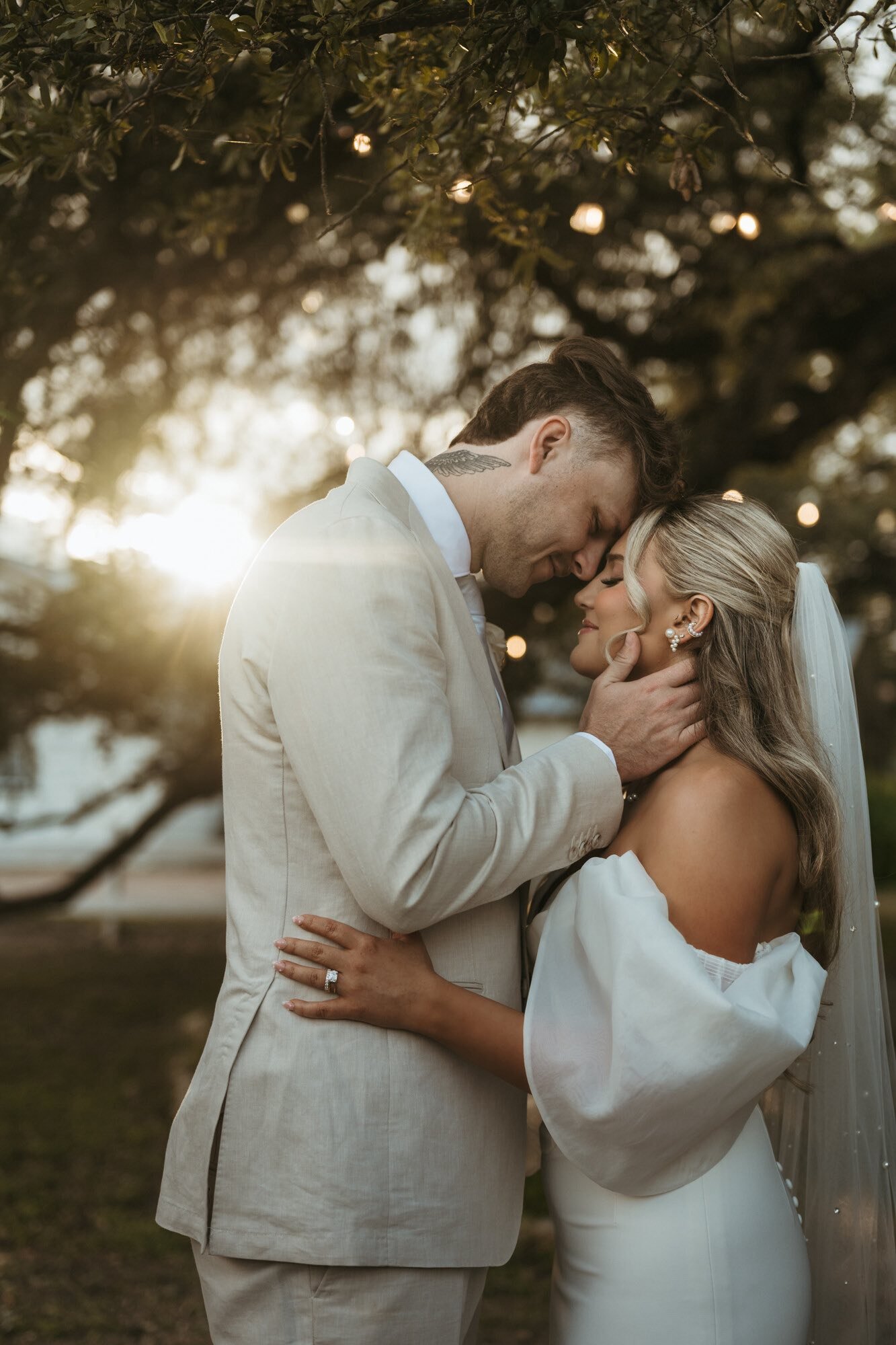 A bride and groom sharing a romantic moment outdoors at sunset, with their foreheads touching and eyes closed, surrounded by trees and warm sunlight.