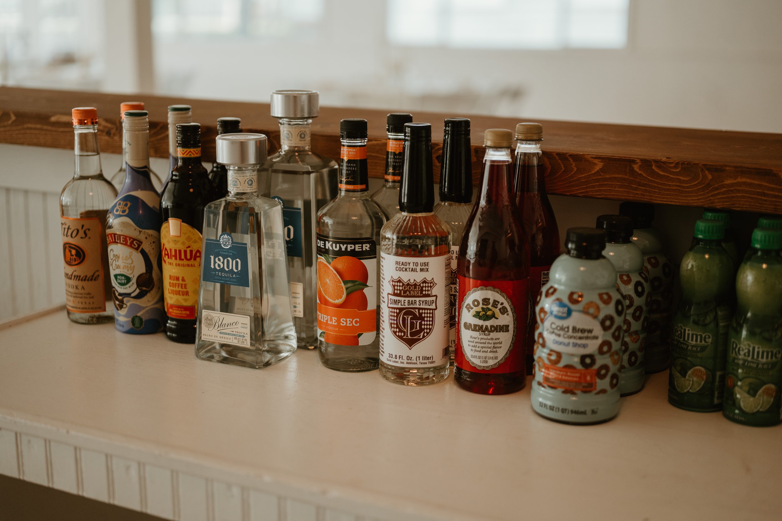 Assorted bottles of alcohol and mixers on a countertop, including tequila, rum, cocktail mix, grenadine, and lemon-lime soda.