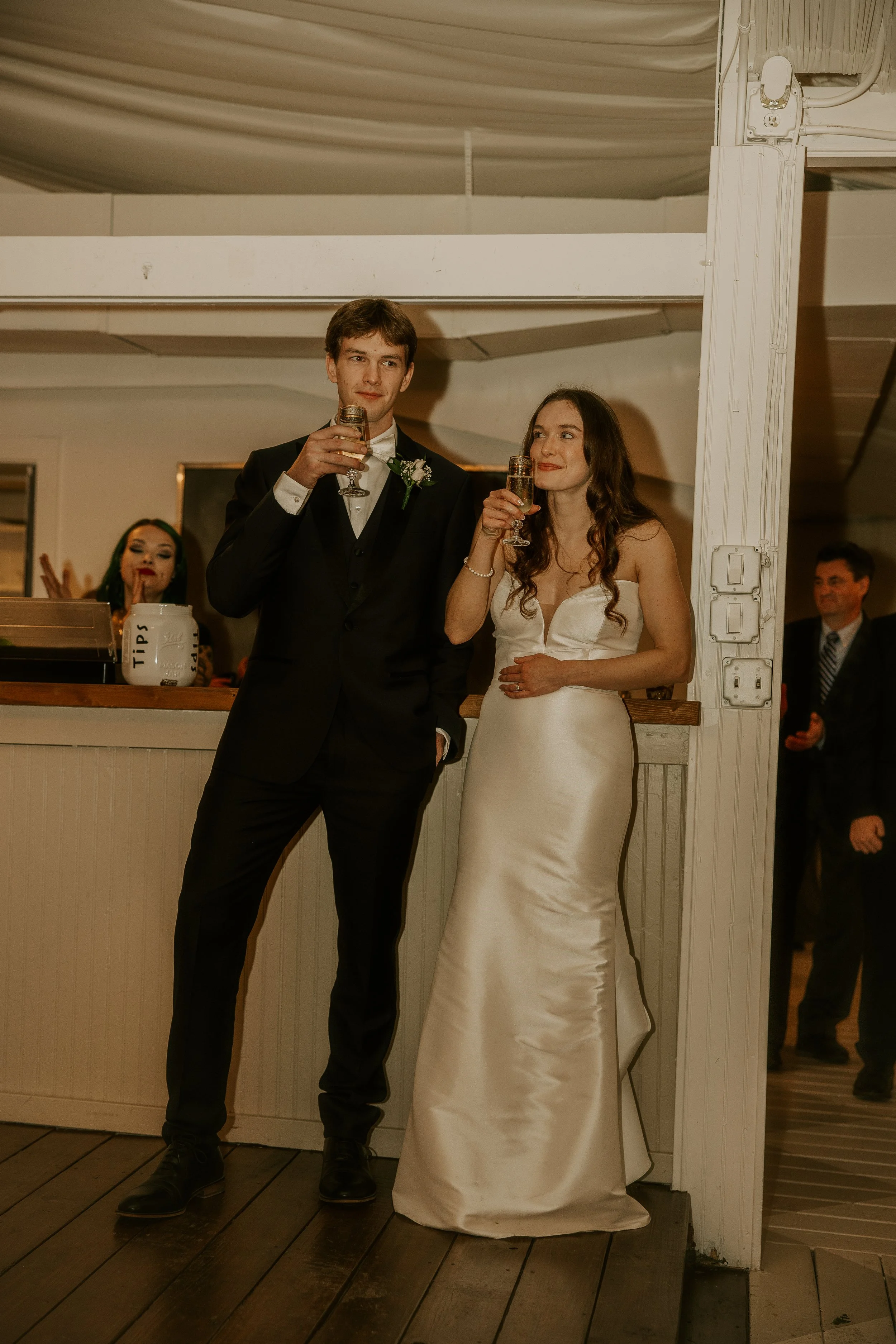 A bride and groom standing together at their wedding reception, holding glasses of champagne, with a bartender and guests in the background.