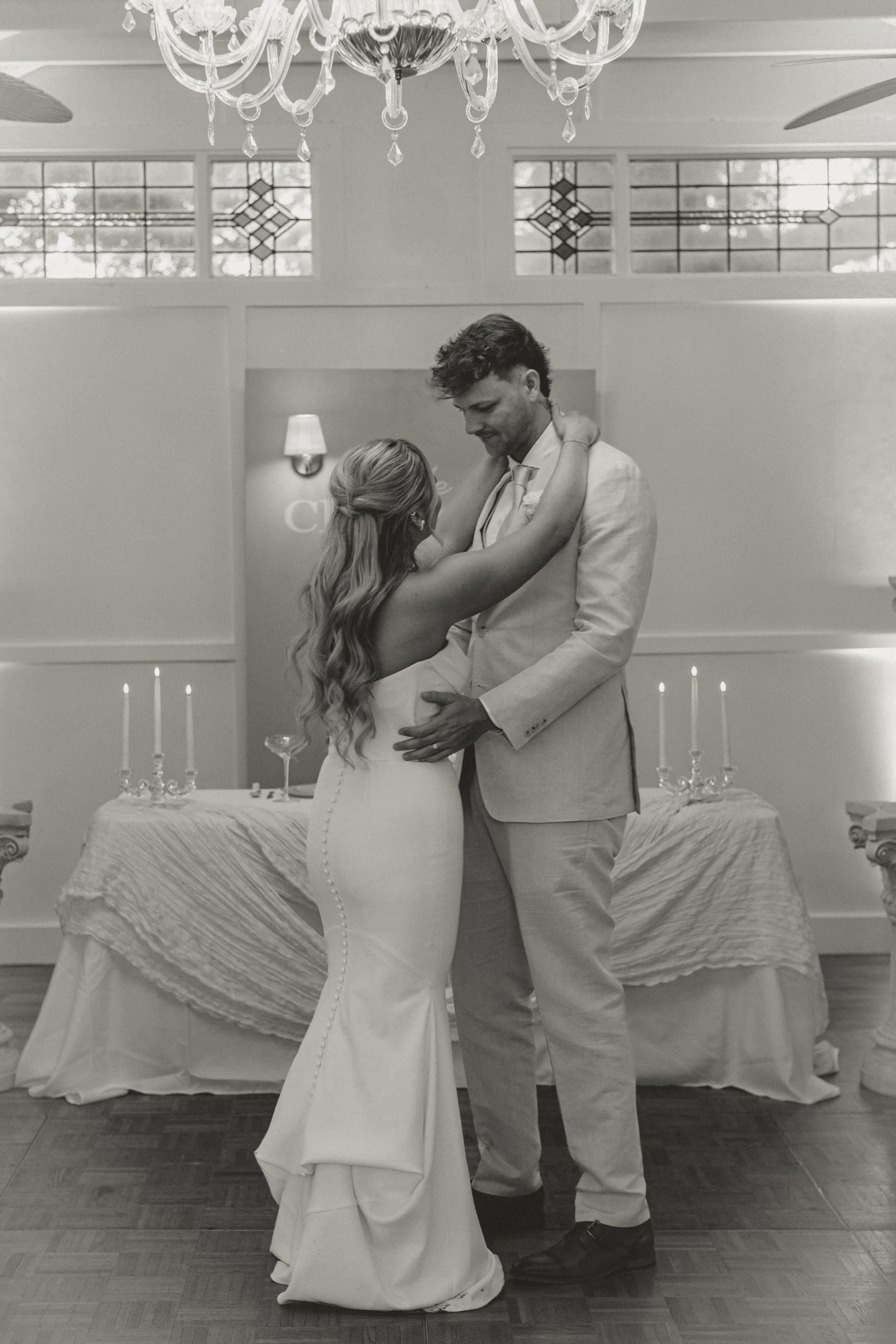 A black-and-white photo of a bride and groom sharing a dance at their wedding reception in a decorated indoor venue with a chandelier, candles, and stained glass windows.