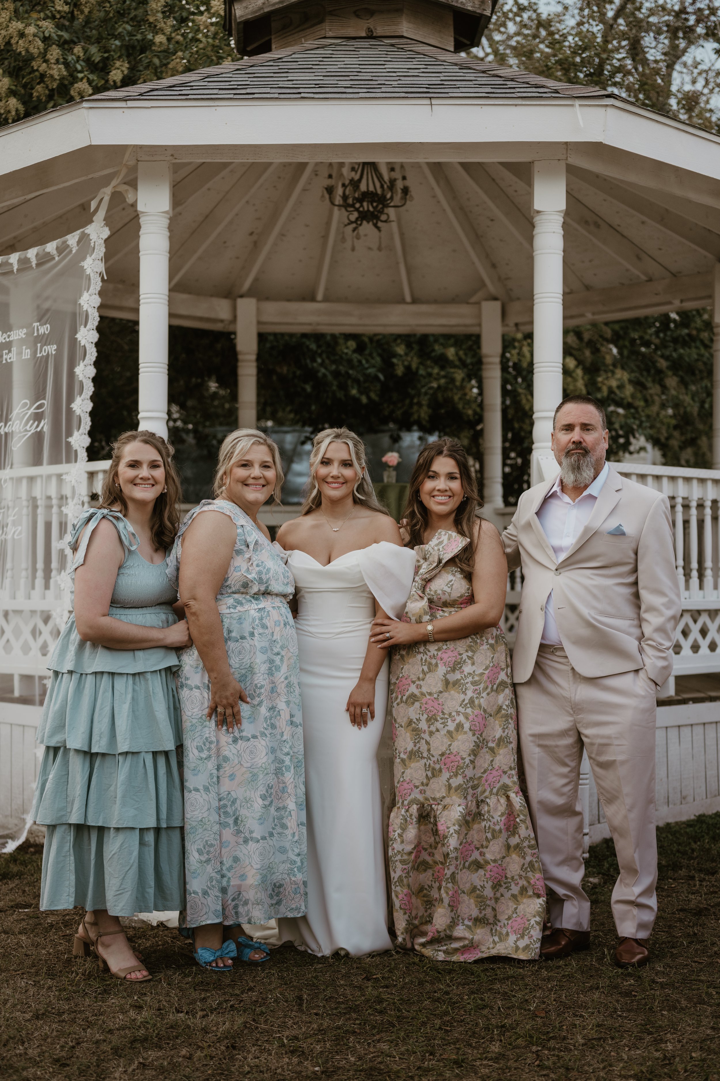 A group of five people standing in front of a white gazebo at an outdoor wedding. The bride is in the center wearing a white off-shoulder wedding dress. Two women are on her left, dressed in floral and light blue dresses, and a man in a cream-colored