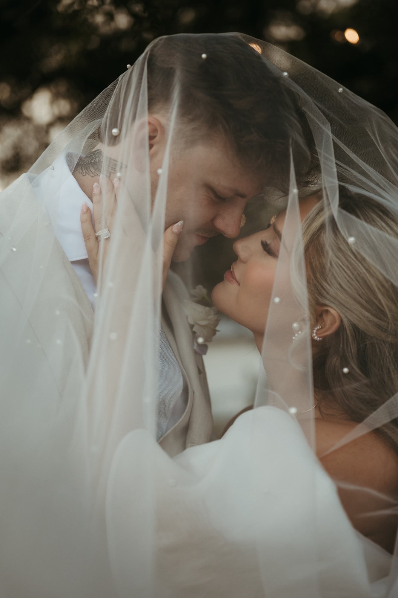 A bride and groom share a close, intimate moment under a sheer veil with pearls, with their foreheads touching and eyes closed, during their wedding.