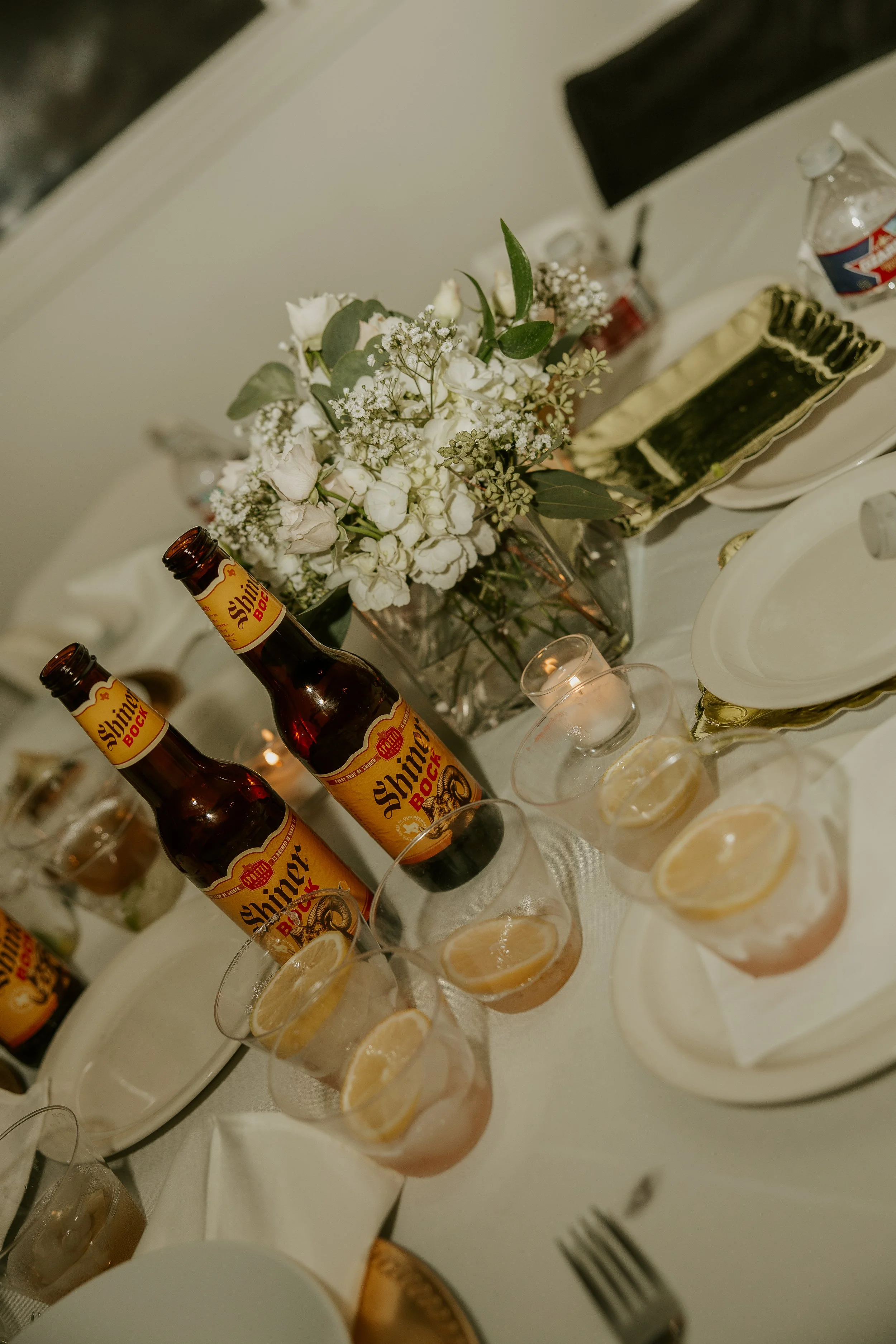 A dining table with bottles of Shiner Bock beer, glasses with lemon slices, a flower arrangement, and plates with appetizers, set for a meal.