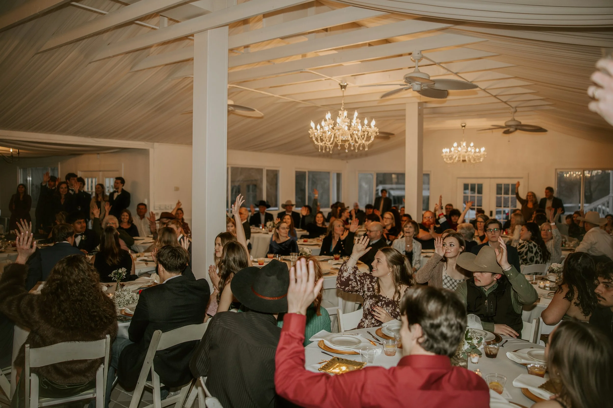 A large indoor gathering of people seated at decorated tables during an event, raising their hands, with chandelier lighting and ceiling fans in the background.