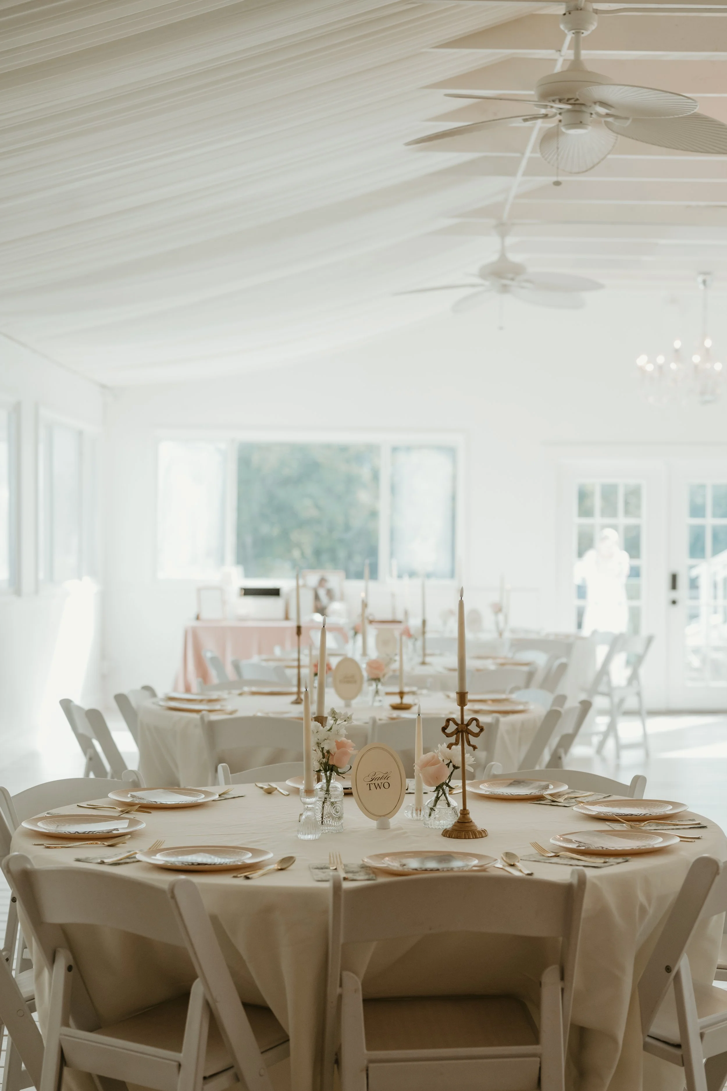 Elegant indoor event space decorated for a celebration with round tables, white chairs, pink and white floral centerpieces, and tall white candles on gold holders, with sunlight streaming through large windows.