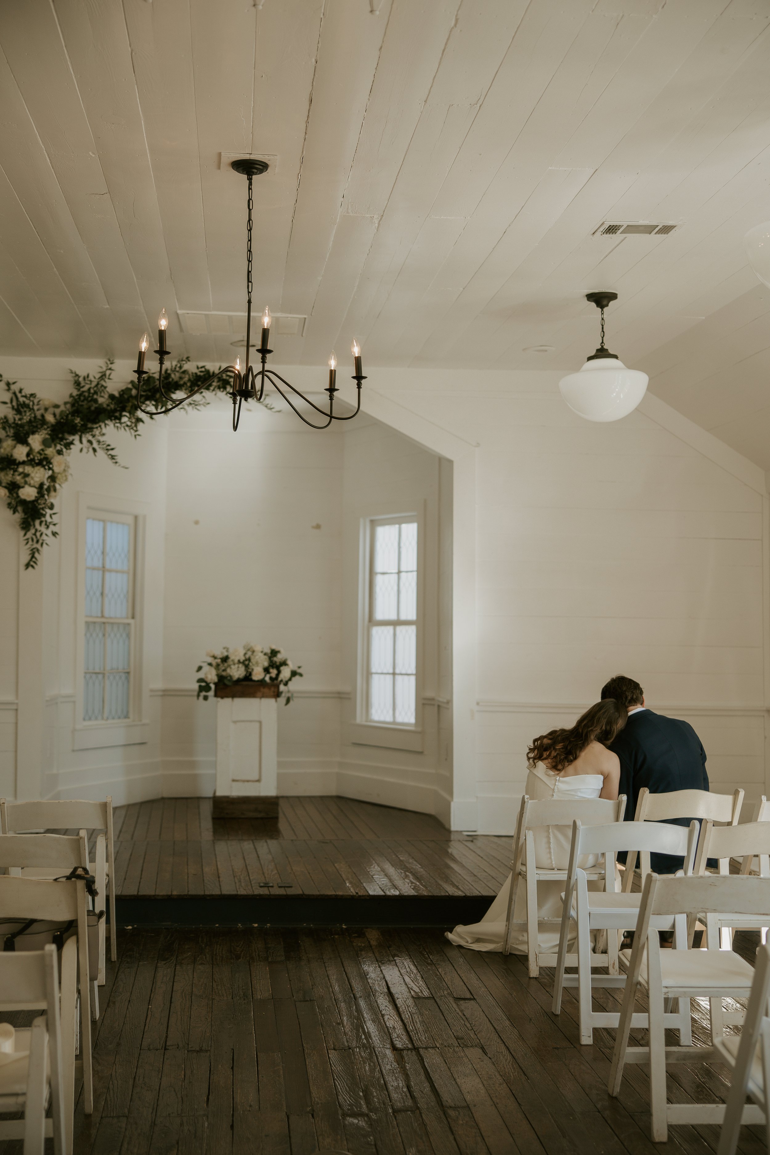 A couple sitting close together on white chairs in a small wedding ceremony space, with an altar decorated with white flowers and greenery, and natural light coming through windows.
