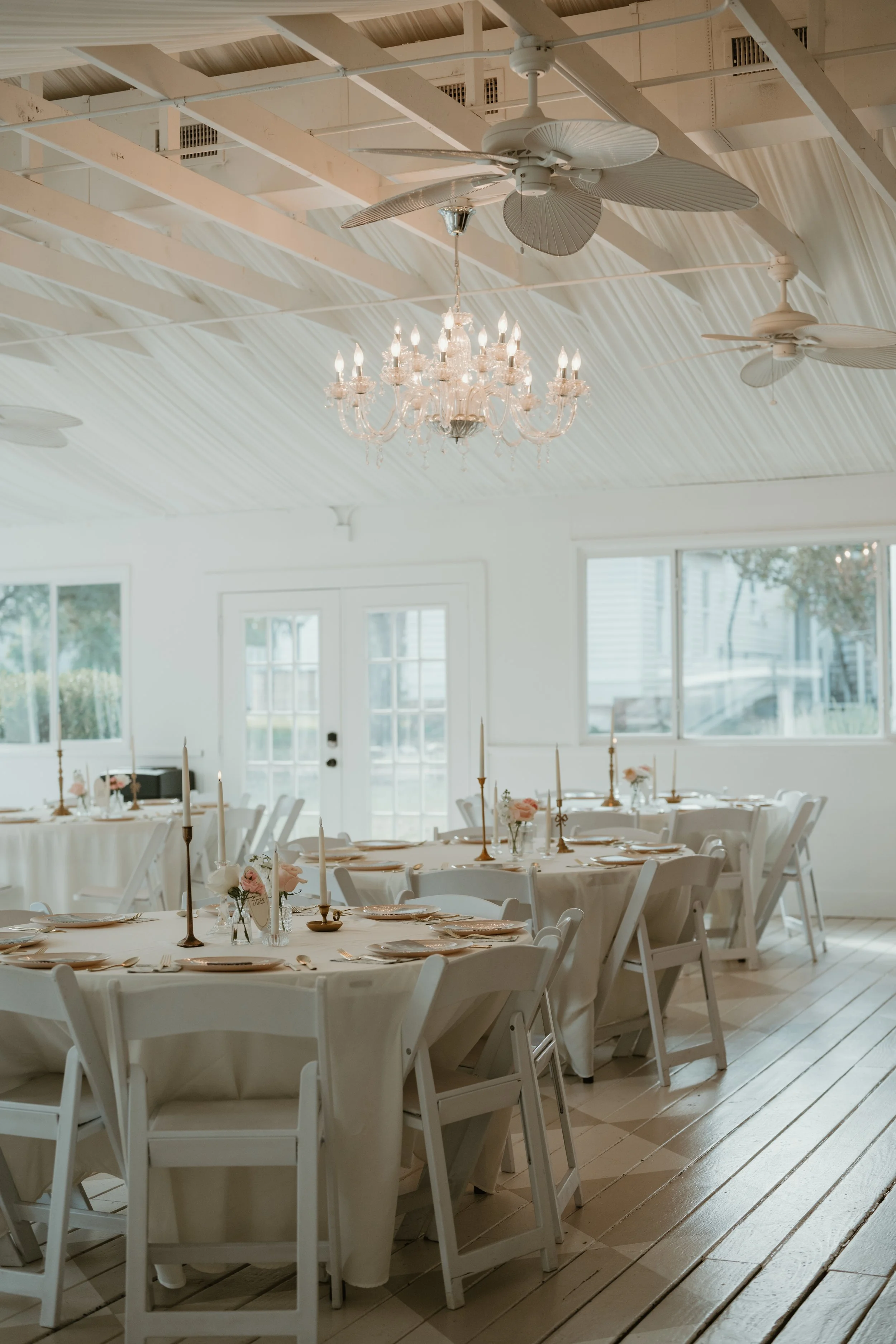 Empty event hall decorated for a formal gathering with round tables covered in white tablecloths, set with plates, cutlery, and floral centerpieces, featuring a chandelier and ceiling fans in a bright room with large windows.