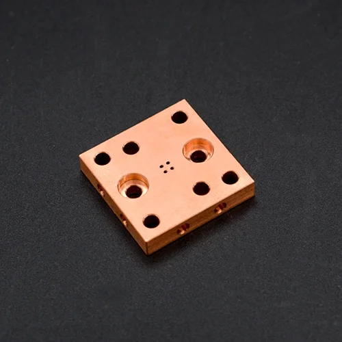 Square copper block with multiple holes on a black surface.