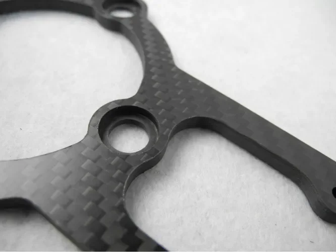 Close-up of a black, textured metal gasket or spacer with circular holes, placed on a white surface.