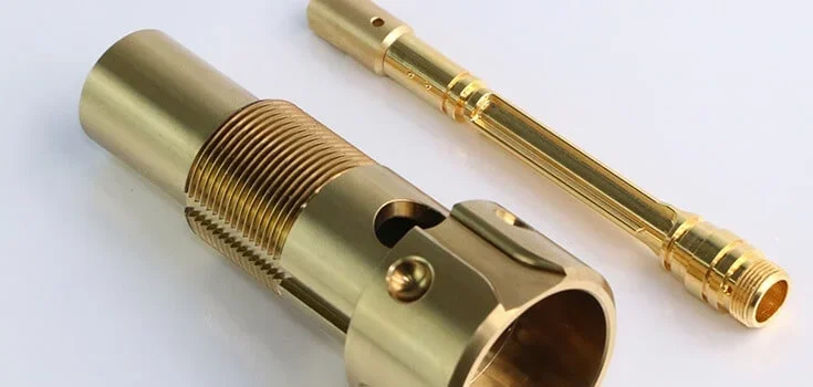 Two gold-colored metal parts, one cylindrical with threaded sections and a slot, and the other slim and elongated with threaded ends.