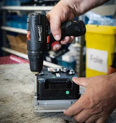 Hands using a cordless drill to tighten a screw on a metal device on a workbench.