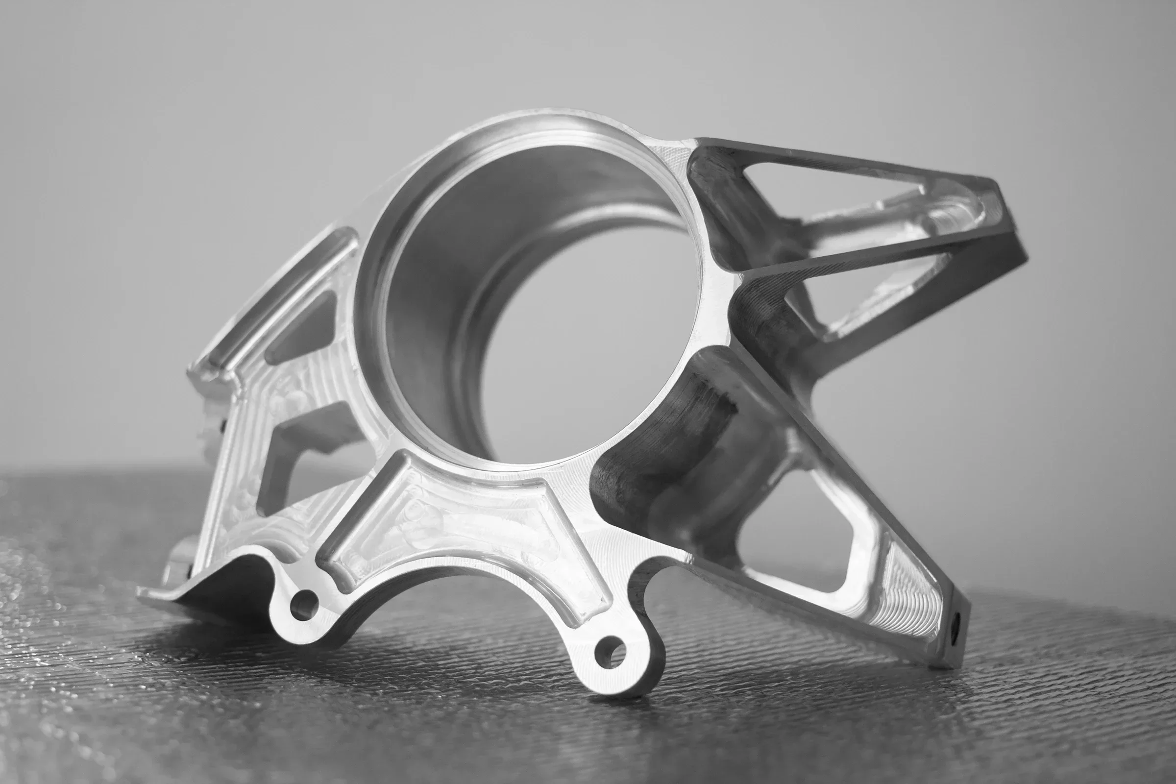 Black and white photo of a metal mechanical part with a cylindrical opening and intricate design, resting on a textured surface.