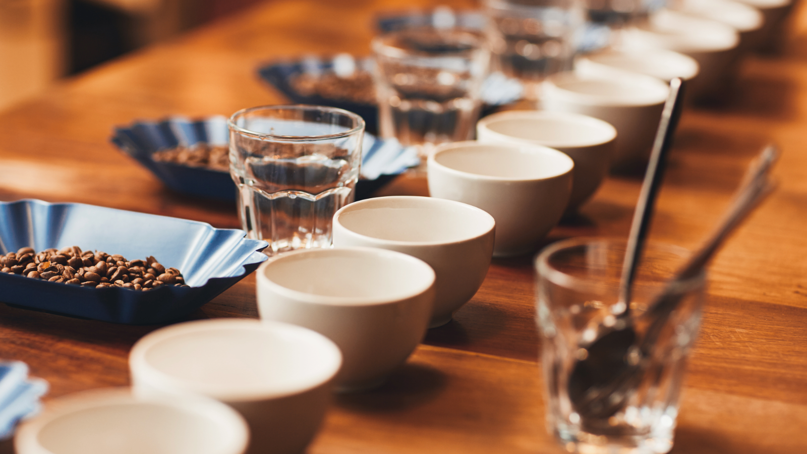 A wooden table set with white bowls, glasses of water, small blue bowls filled with coffee beans, and glasses with spoons, arranged in a line, warm natural lighting.