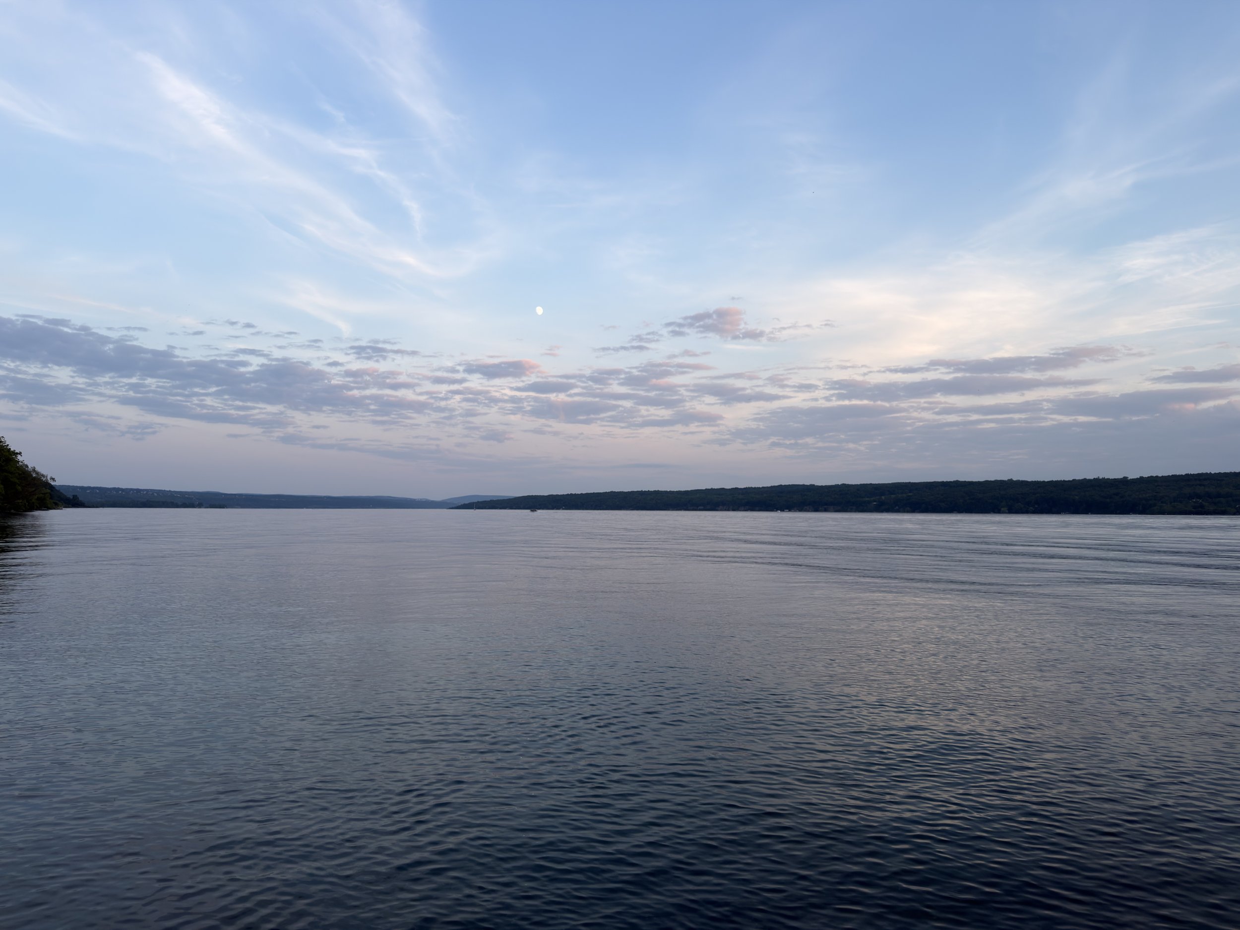 A wide river or lake under a blue sky with scattered clouds. The horizon shows a distant shoreline with hills or forests.