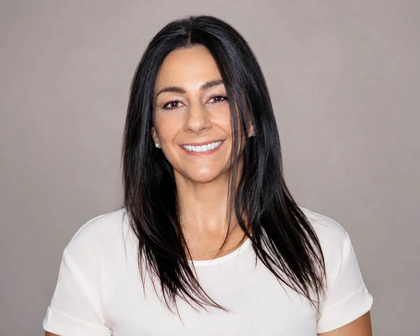 A woman with long straight black hair, smiling, dressed in a white top, against a plain gray background.