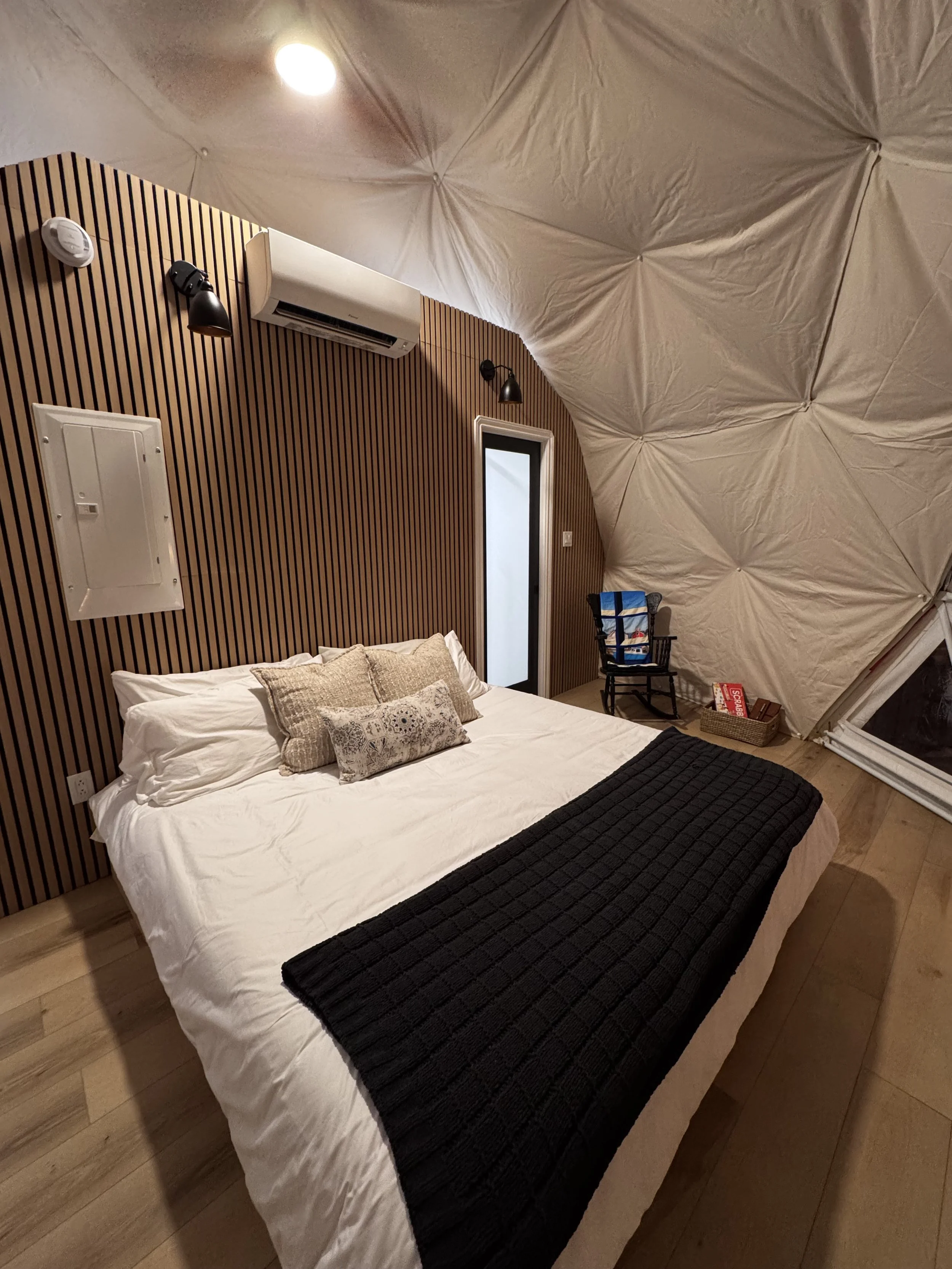 A bedroom with a bed covered in white and black bedding, against a wooden slat wall. The ceiling is curved and covered with beige fabric, illuminated by recessed lighting and wall-mounted lamps. There is a small door, a chair with a bag on it, and a basket with guests' items nearby.