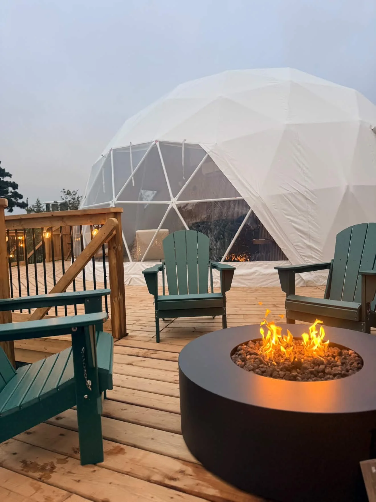 A wooden outdoor deck with Adirondack chairs and a fire pit, with a modern geodesic dome structure in the background and cloudy sky.