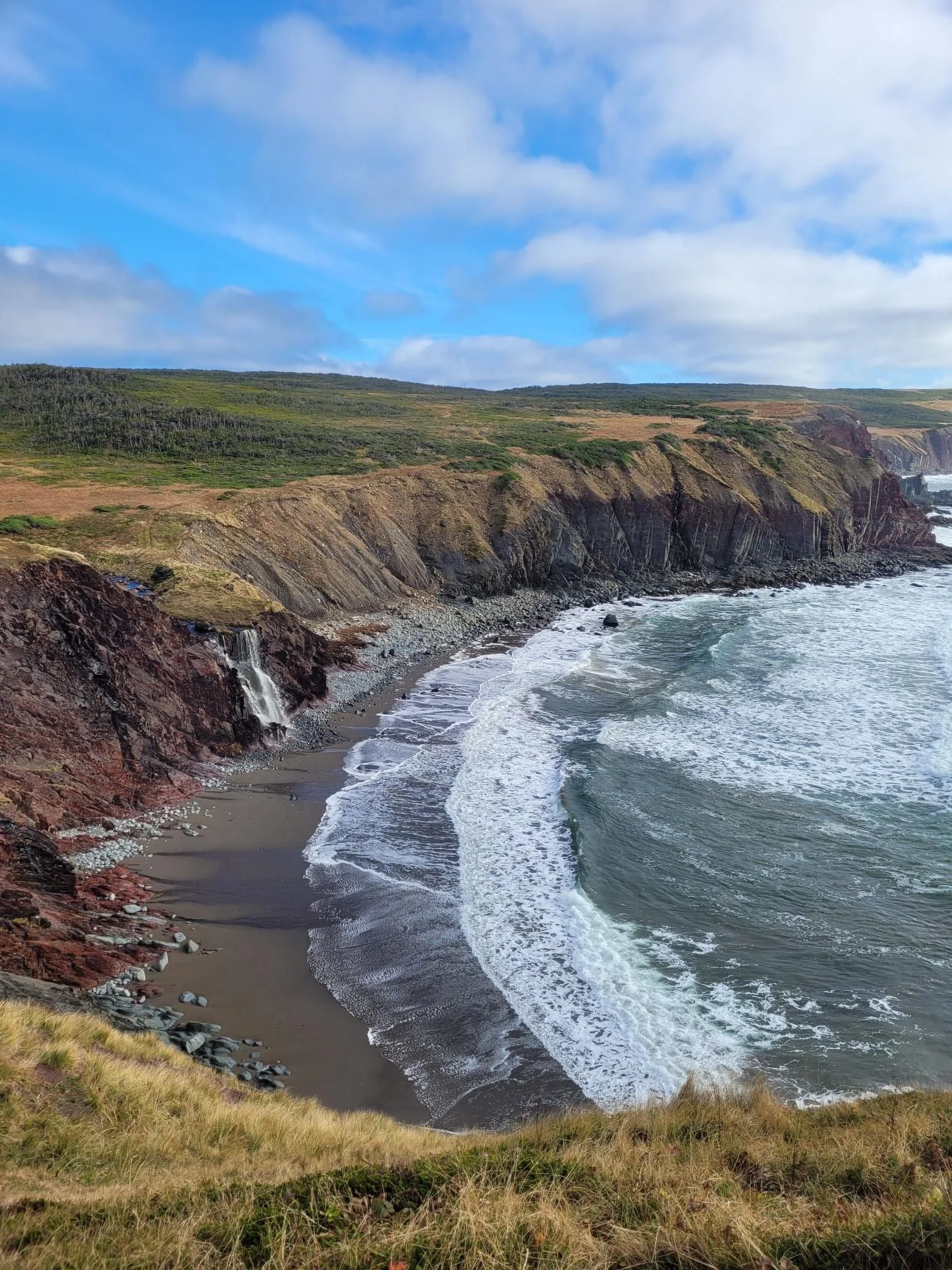 Coastal cliffs with a small waterfall flowing onto a sandy beach, choppy ocean waves, green grassy top, partly cloudy sky. This is called red land, located near Cape St. Mary's