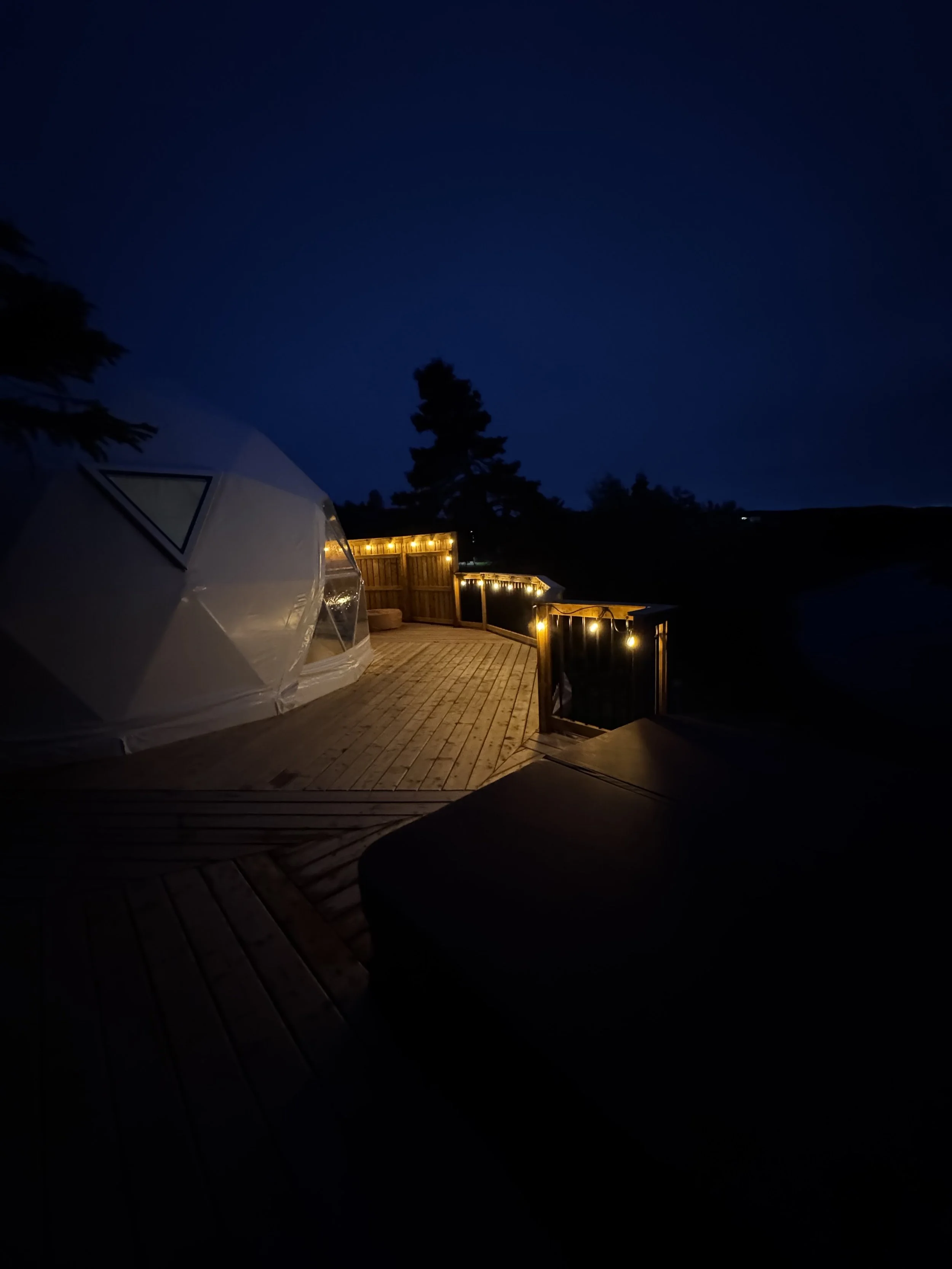 A nighttime scene of a wooden deck with string lights, a fence, a small white geodesic dome, and trees in the background under a dark sky.
