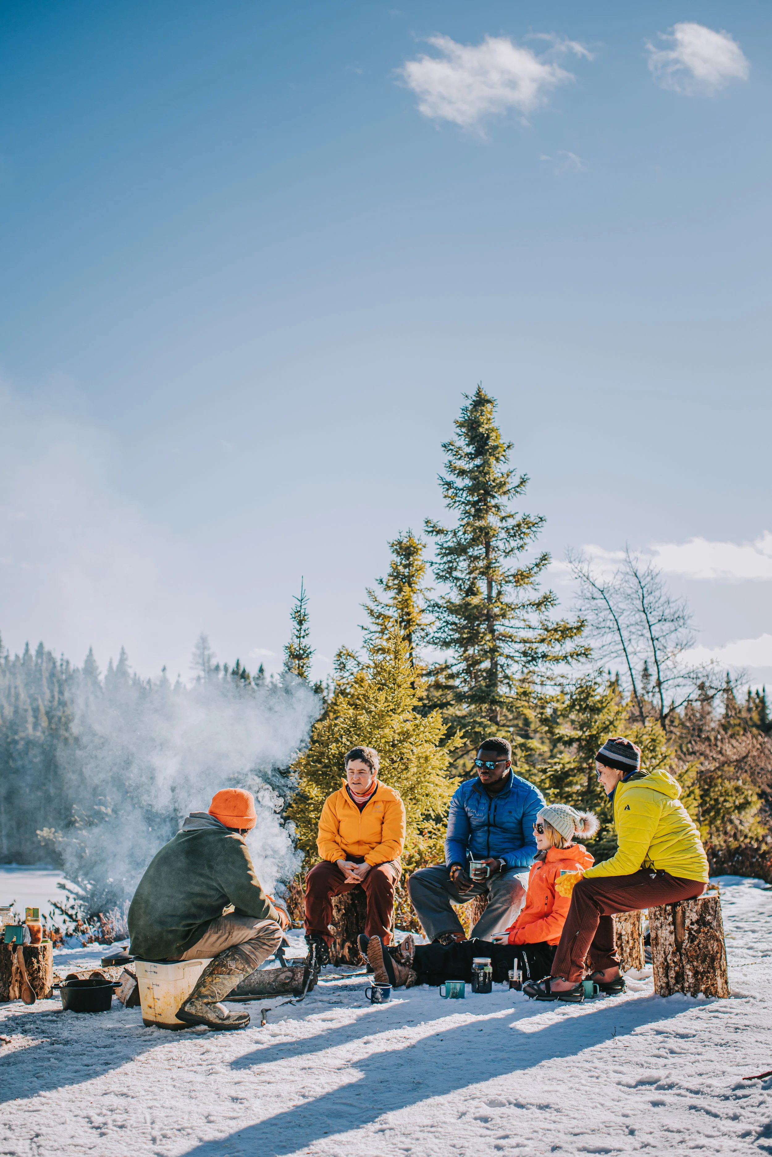 A group of people gathered around a campfire outdoors on snow-covered ground, with evergreen trees and a clear blue sky in the background.