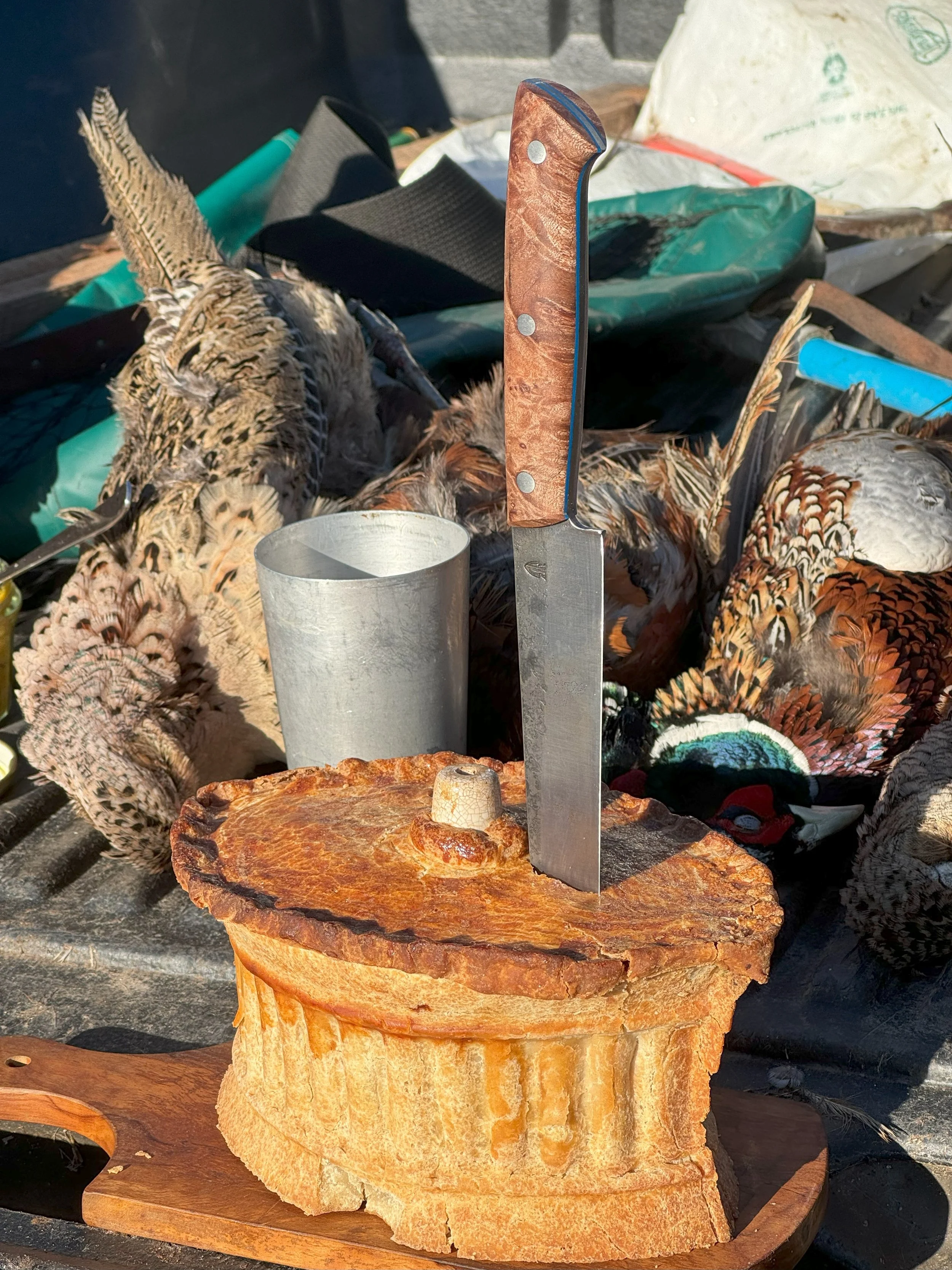 A hunting knife with a wooden handle and a metal blade stands upright on a round, polished stone surface. The background shows dead pheasants, a metal cup, and various outdoor items.