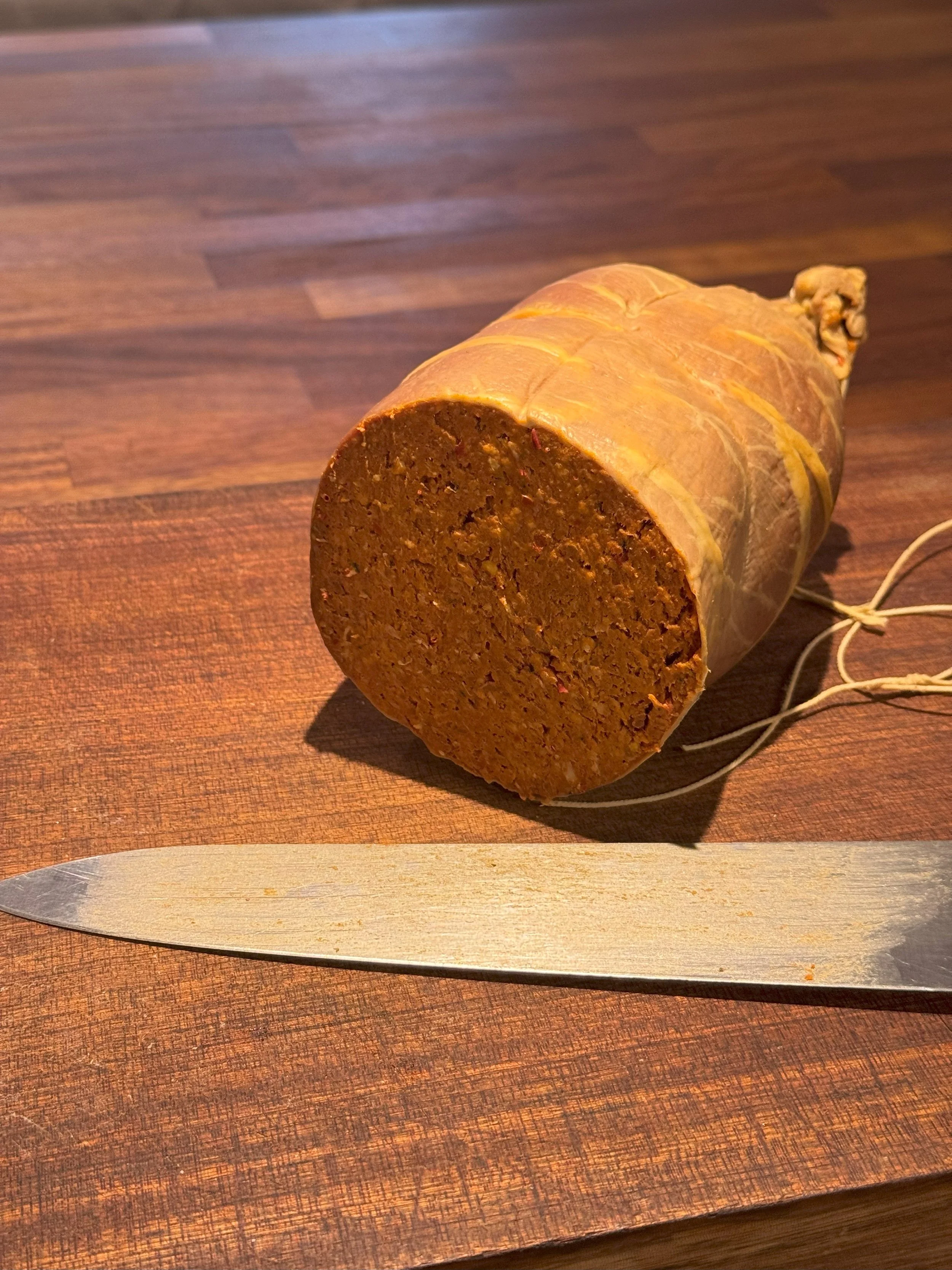 A large loaf of meatloaf with a golden-brown crust on a wooden table, with a kitchen knife placed in front of it.