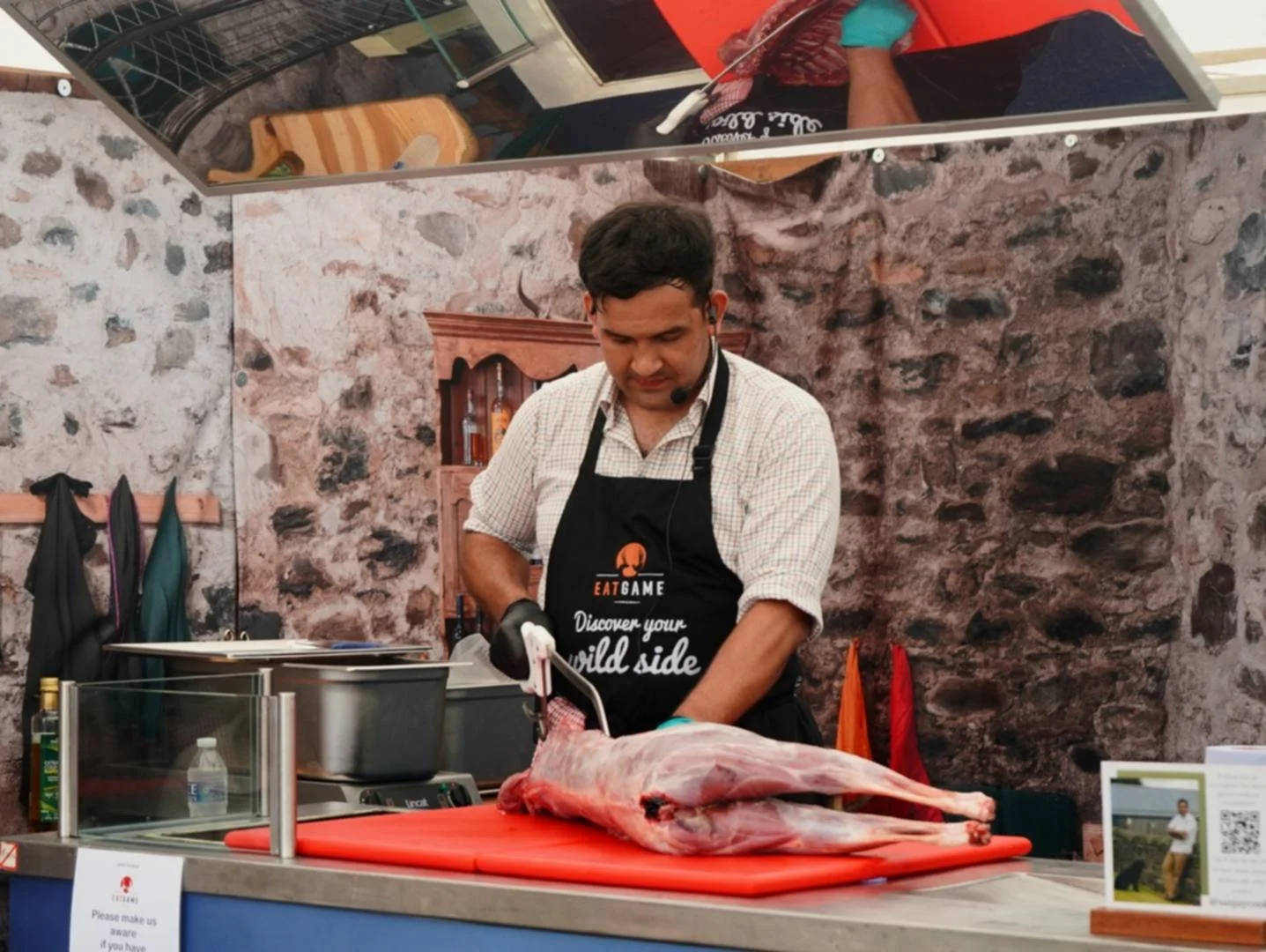 Man in apron preparing a large piece of meat at a cooking demonstration. Stone wall background, red cutting board, and kitchen utensils visible.