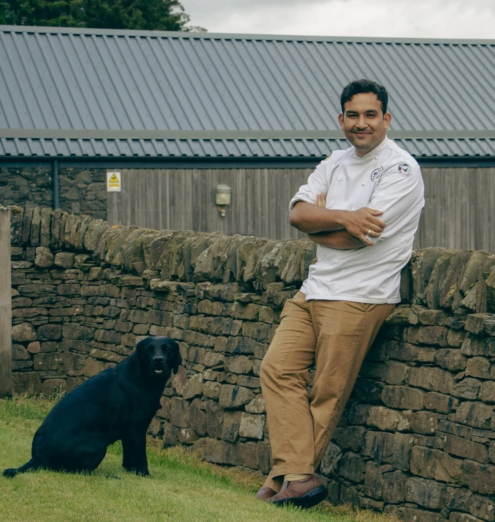 A man in a chef's coat stands outdoors in front of a stone wall, with a black dog sitting on the grass in front of him. The man is smiling with arms crossed, and the dog has its tongue out, looking toward the camera.