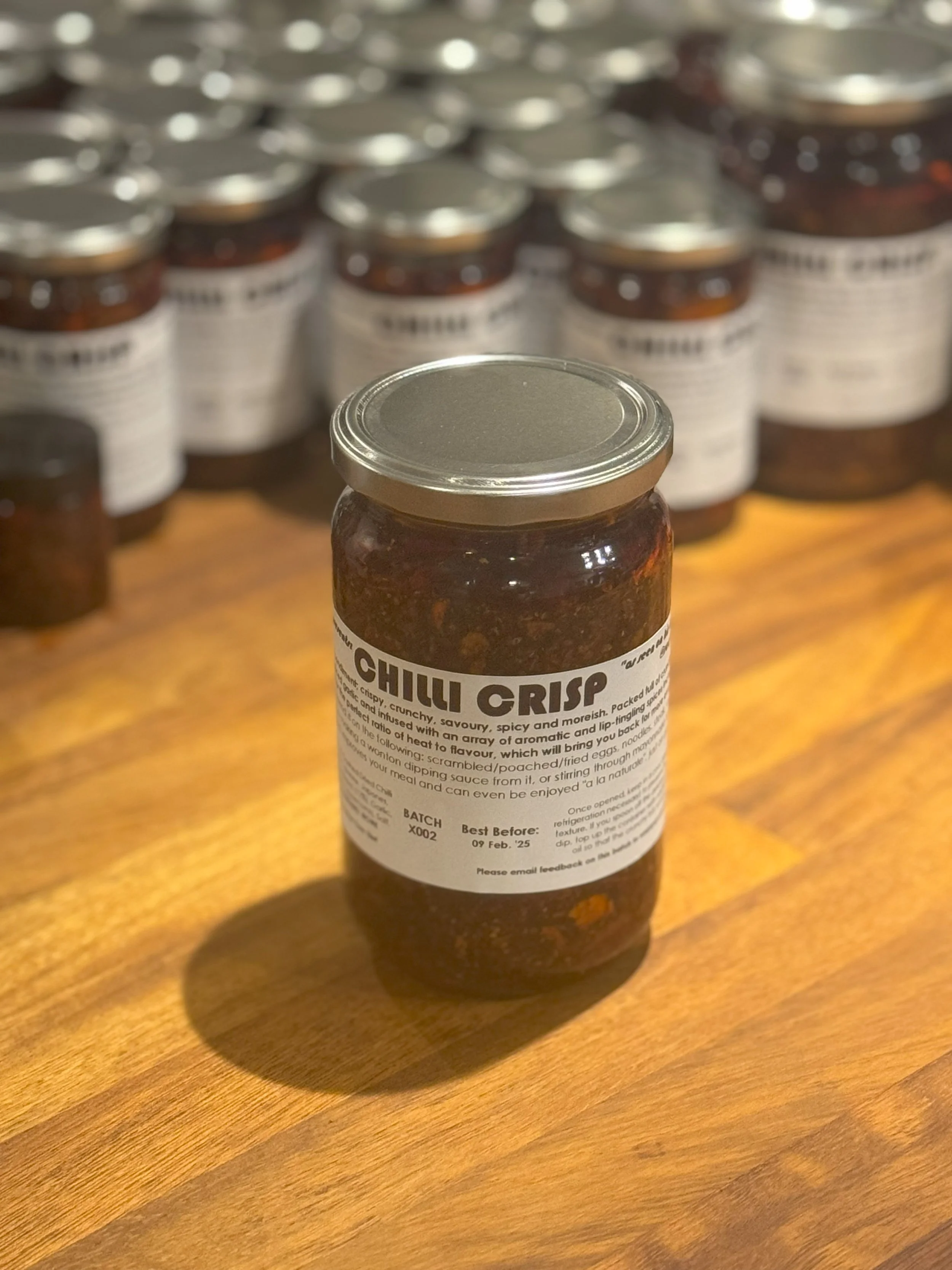 A bottle labeled 'Chilli Crisp' placed on a wooden surface with numerous similar bottles in the background.