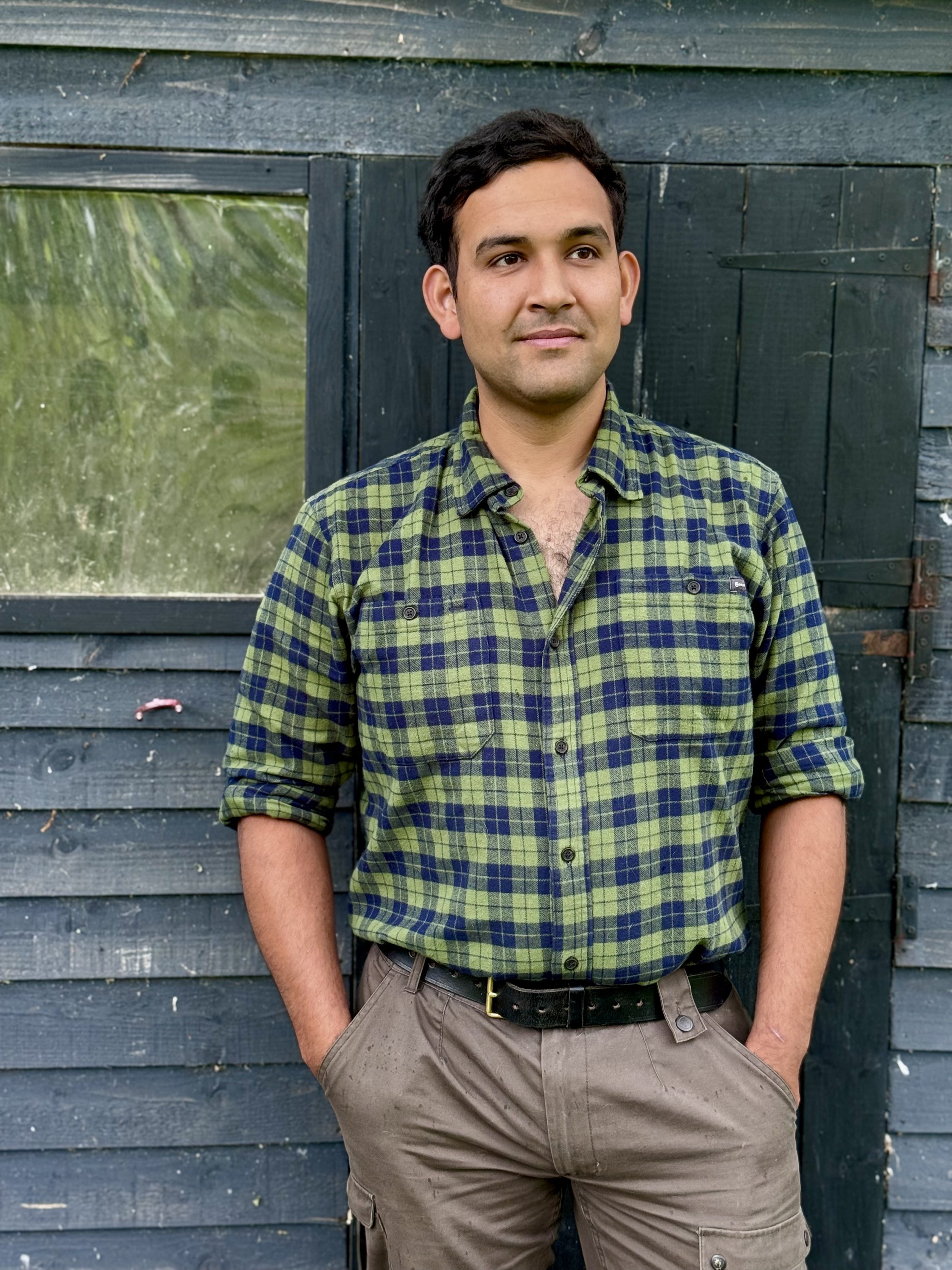 A man with short dark hair wearing a green plaid shirt with rolled-up sleeves and gray cargo pants standing in front of a weathered black wooden wall.