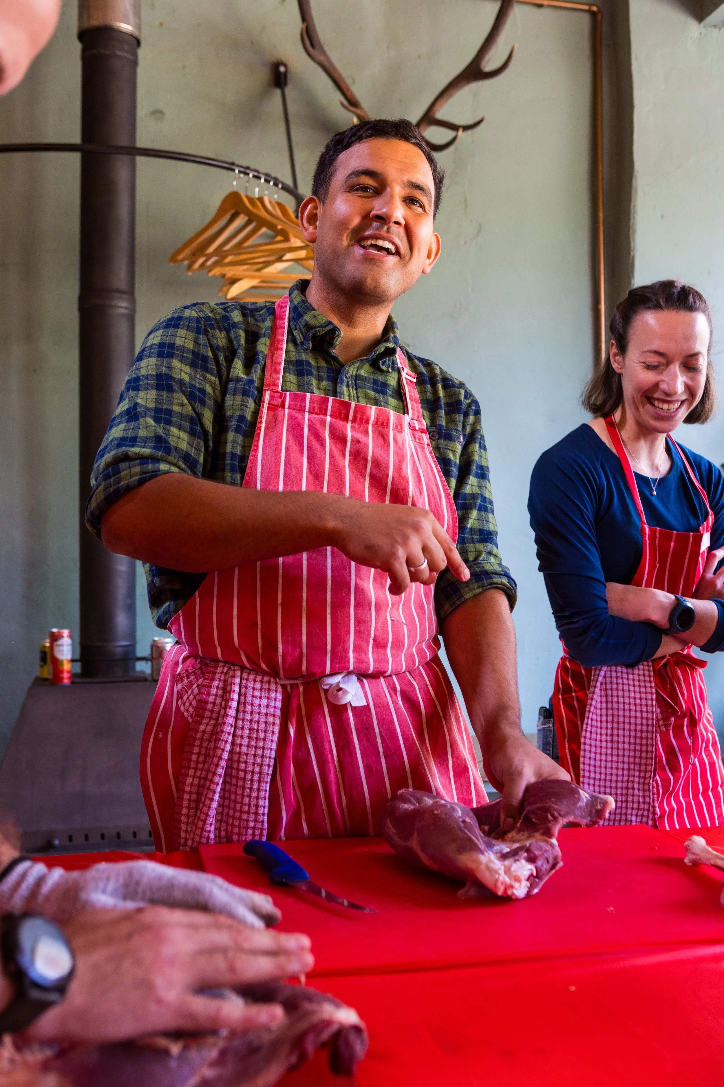 Two people wearing red and white striped aprons preparing meat, with one man smiling and a woman laughing in a rustic kitchen setting.