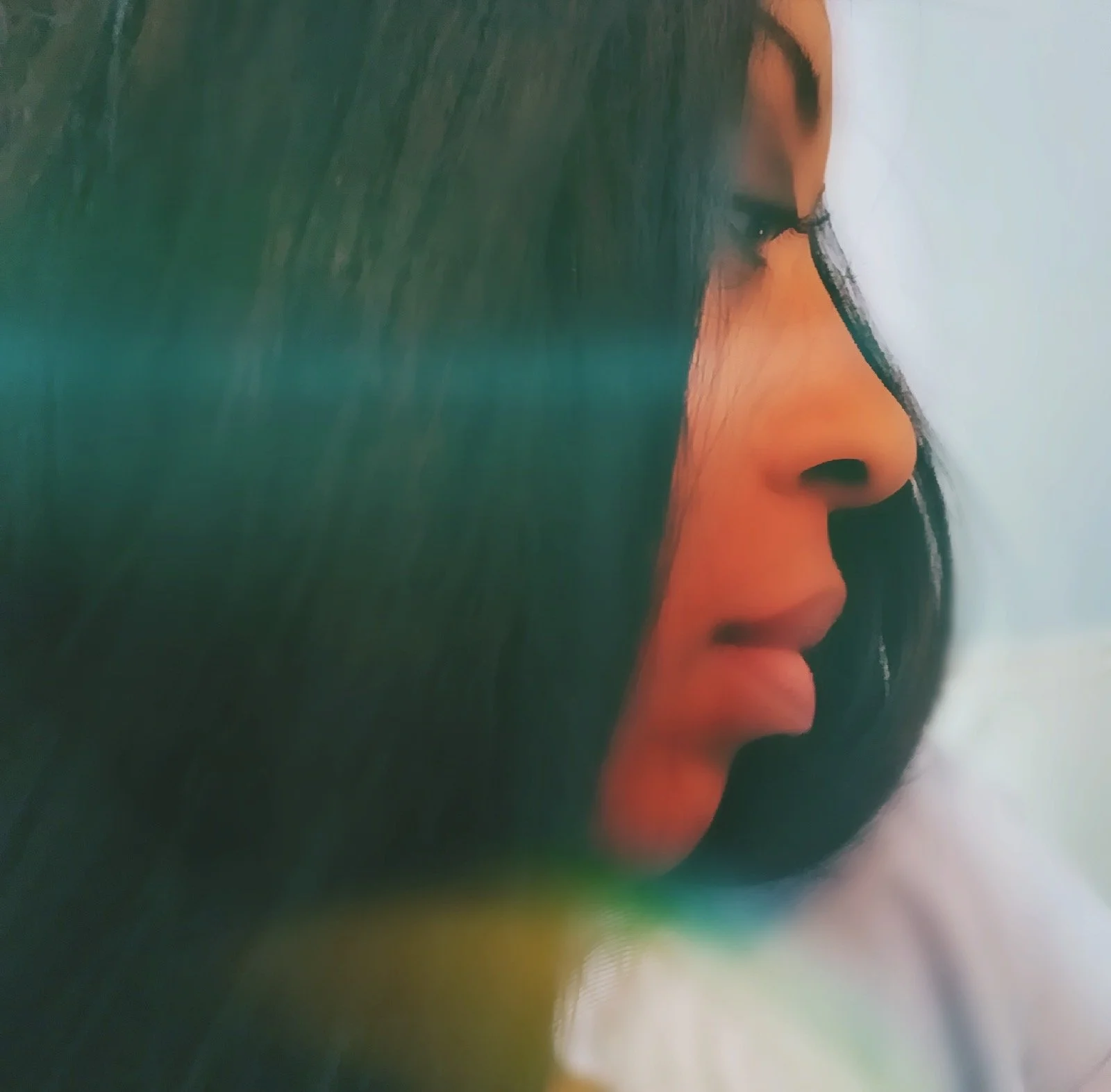 Close-up profile of a woman with long dark hair, showing her nose, lips, and closed eyes, with soft lighting and a blurred background.