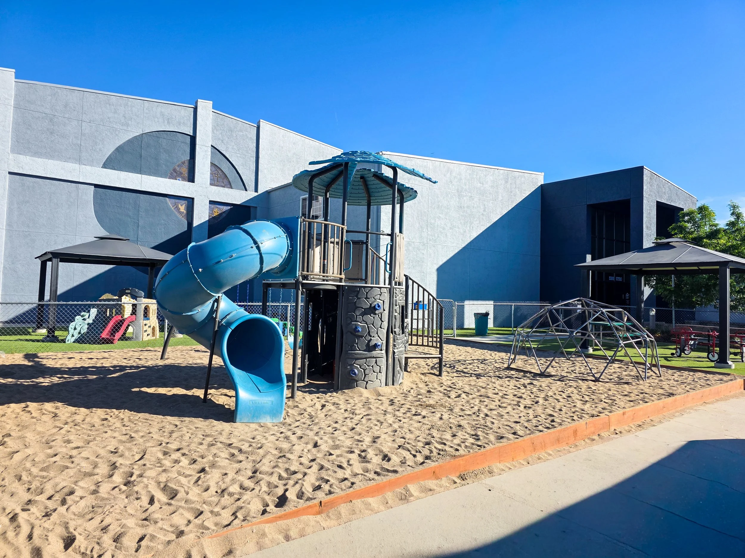 Playground structures including a tube slide, a playhouse, and a climbing wall, with a scenic mountainous background and cloudy sky.