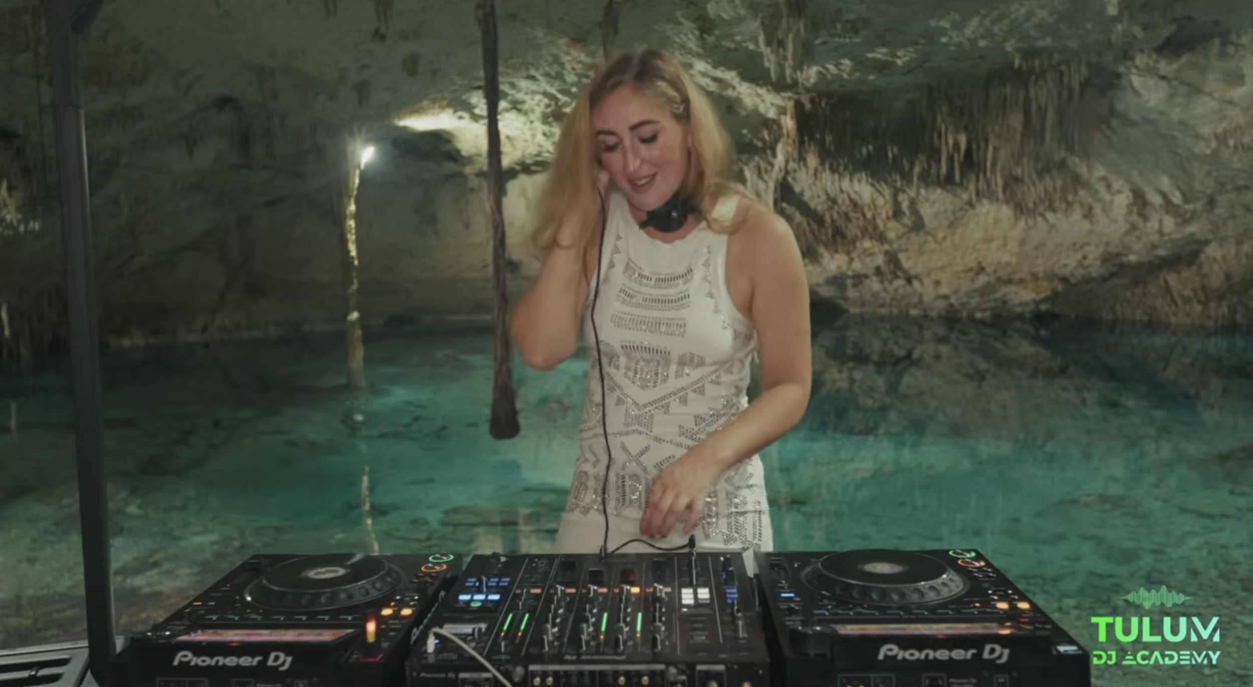 A woman with blonde hair in a rocky cave, DJing with Pioneer DJ equipment, wearing a sleeveless white dress, smiling while looking down. Tulum DJ 