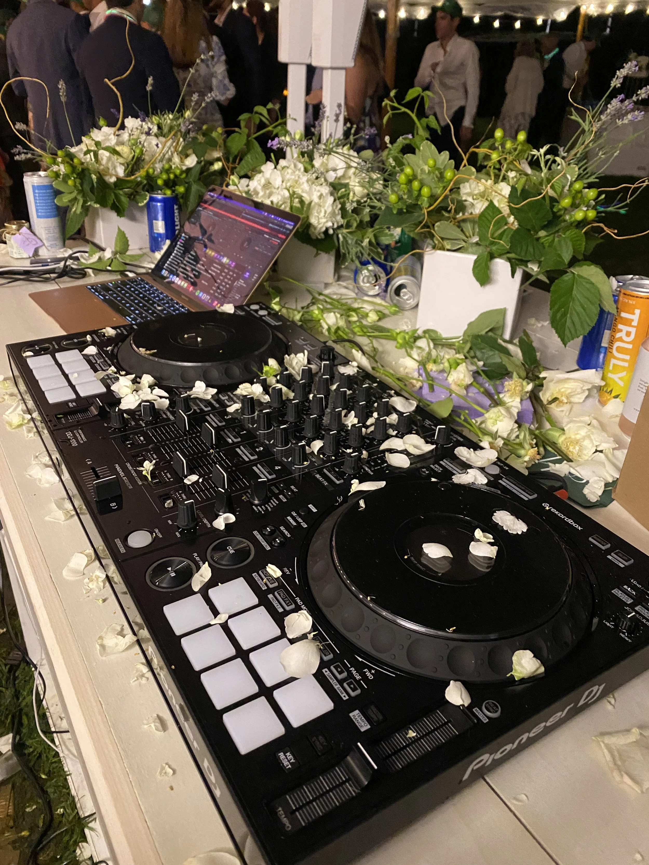 A DJ setup with a Pioneer DJ controller covered in white flower petals, surrounded by floral arrangements and snacks, with a laptop displaying a DJ software interface in the background, at an outdoor event.