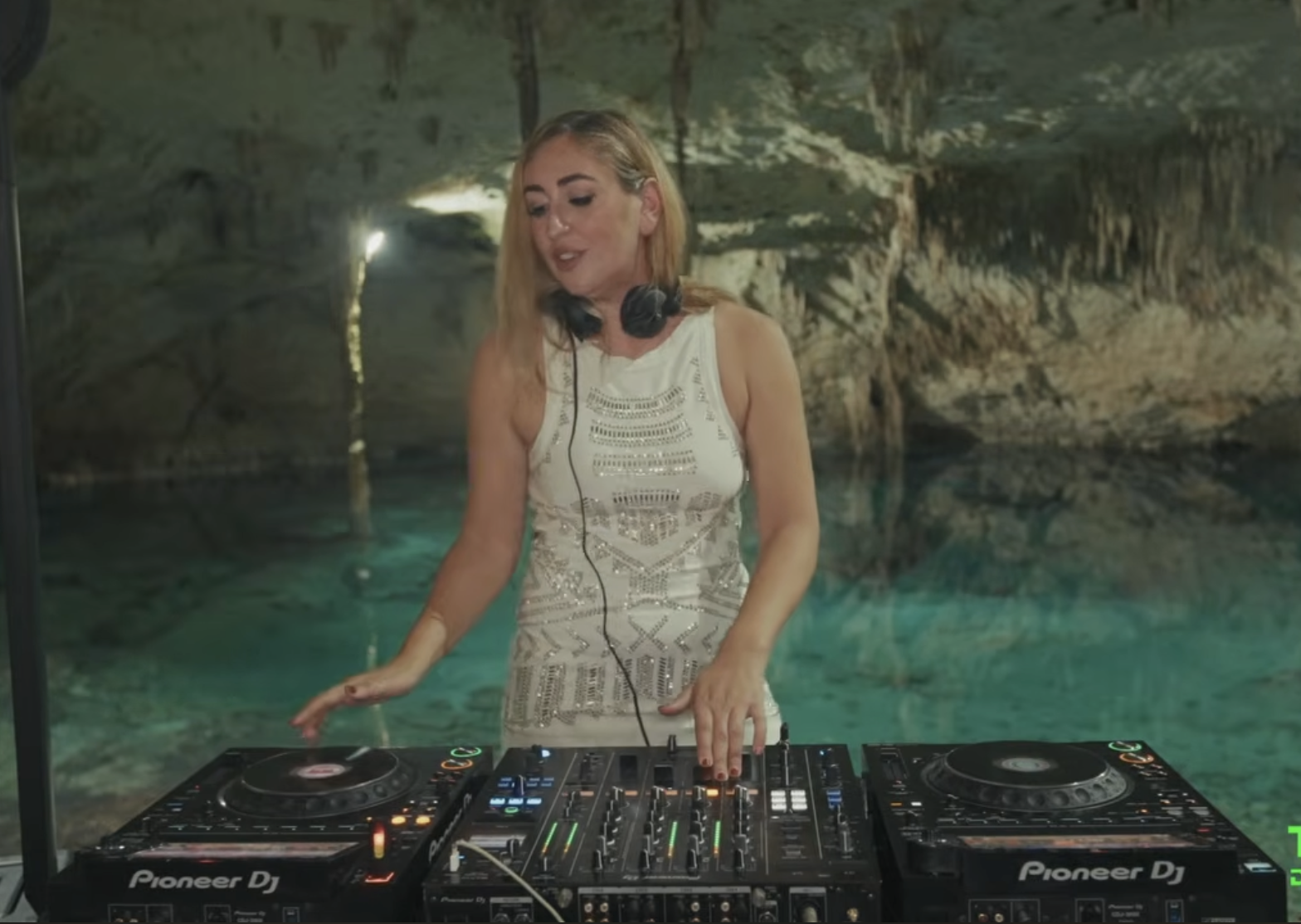 A woman DJ performs in a cavern with turquoise water and rocky walls, wearing a sleeveless white dress and headphones.