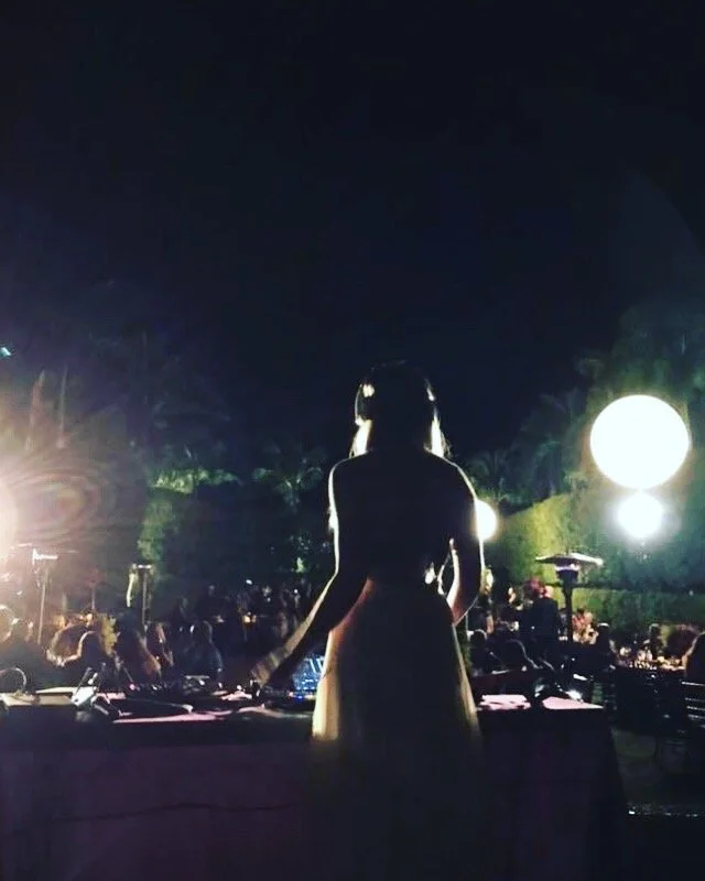 Silhouette of a woman standing behind a DJ setup at an outdoor night event, with bright lights and a crowd in the background.