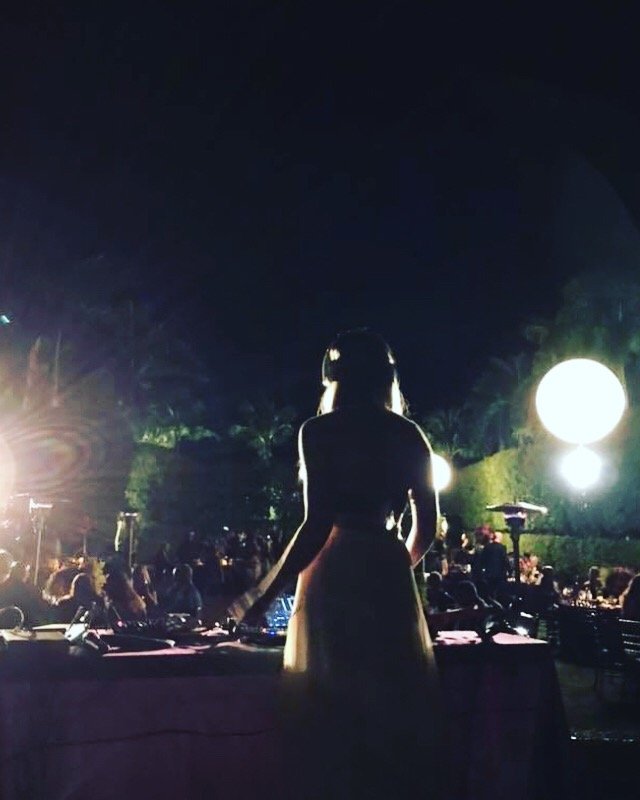 Silhouette of a woman standing at a DJ booth during an outdoor night event, with bright lights and a crowd in the background.