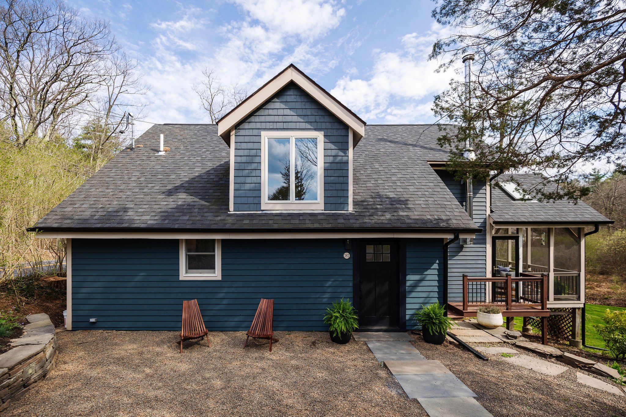 Back view of a blue house with a gable window, surrounded by trees, with a gravel yard and wooden chairs in the foreground.