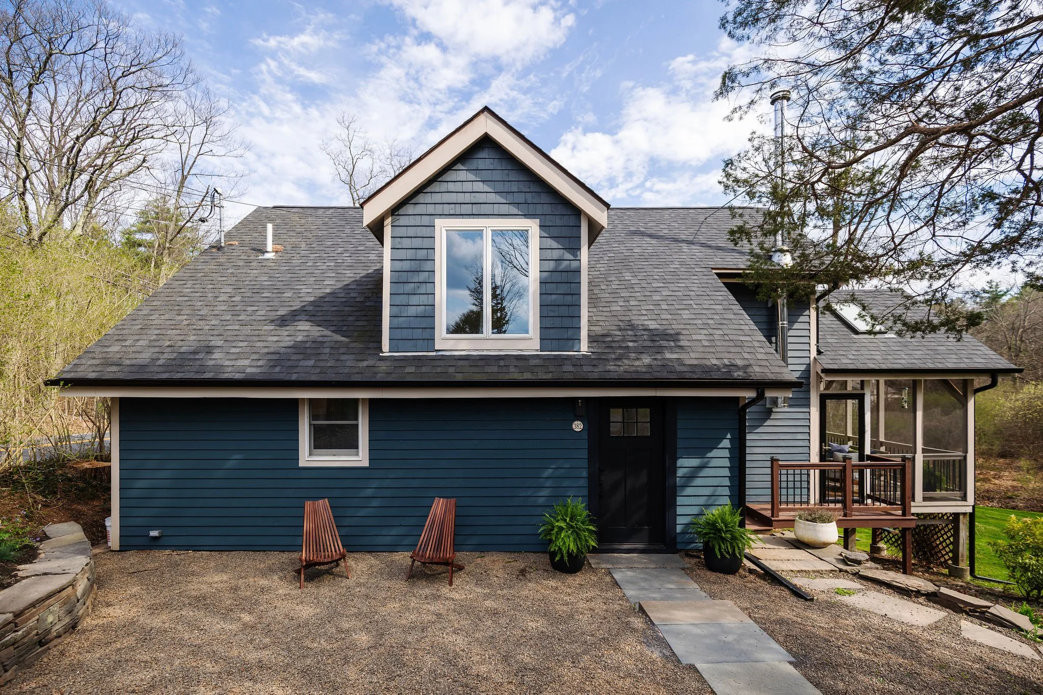 Blue house with dormer window on gray shingle roof, two leaning wooden chairs in front, landscaped yard with small plants, trees, and a wooden deck on the right, under a partly cloudy sky.
