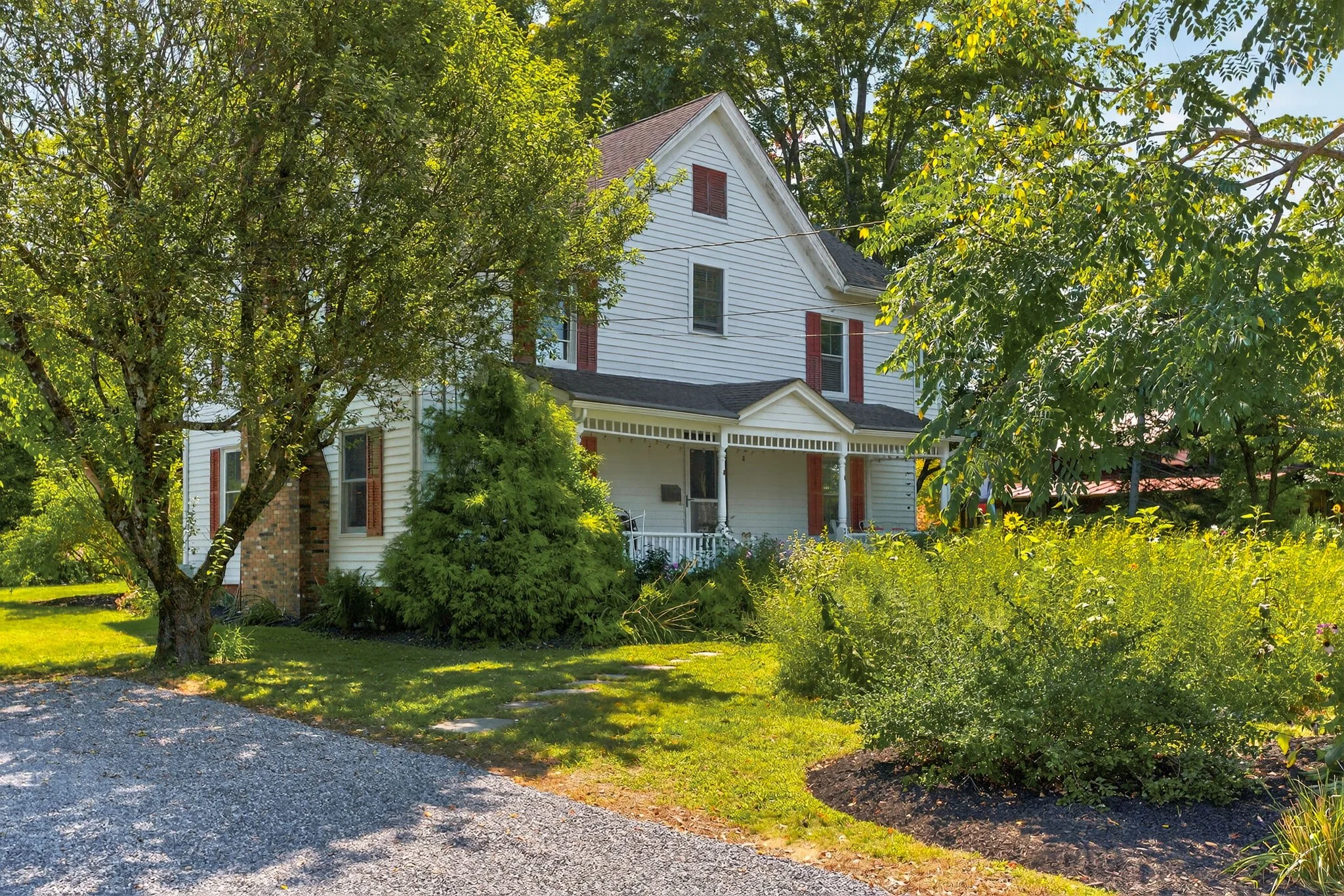 A white two-story house with red shutters, surrounded by green trees and a garden, with a gravel driveway in the foreground.